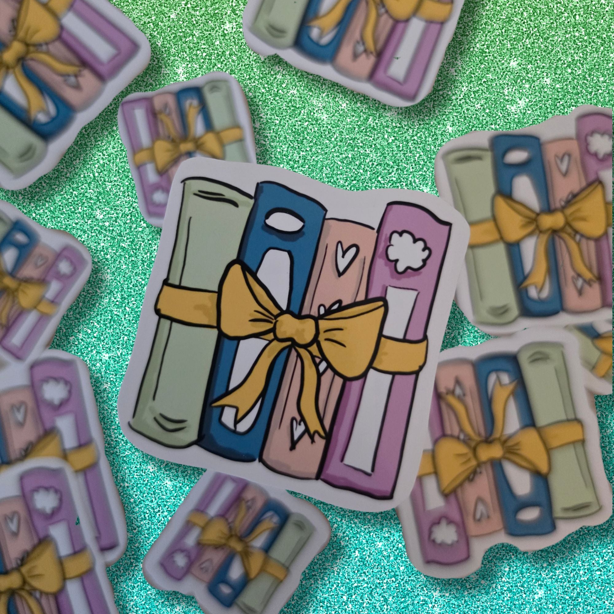 A die-cut sticker featuring a cartoon illustration of colorful books tied with a yellow ribbon and bow, displayed on a sparkling mint green background with other similar stickers scattered around.