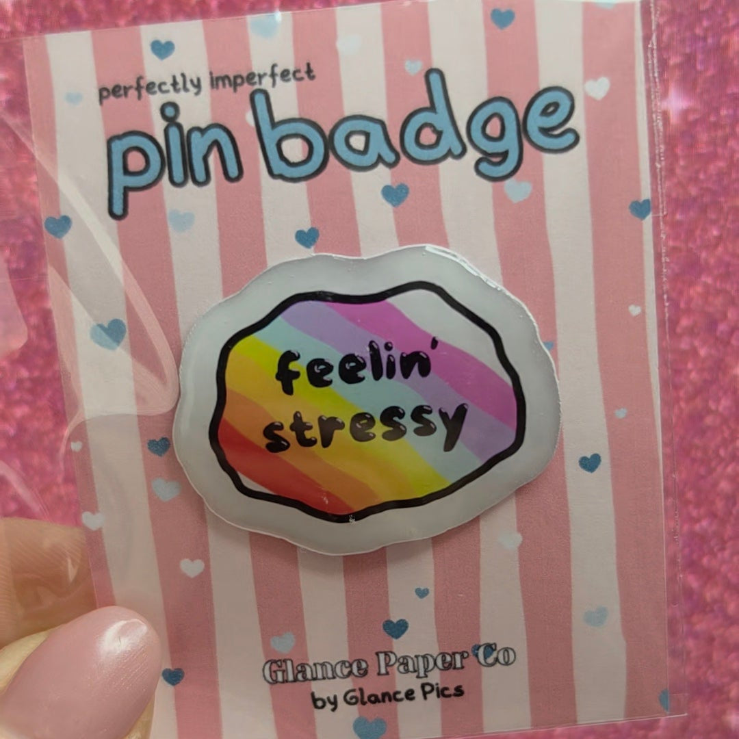 A hand holding a pink and white striped card with a colorful  resin pin badge that reads 'feelin' stressy'. The card is branded 'perfectly imperfect pin badge' and 'Chance Paper Co by Glance Pics
