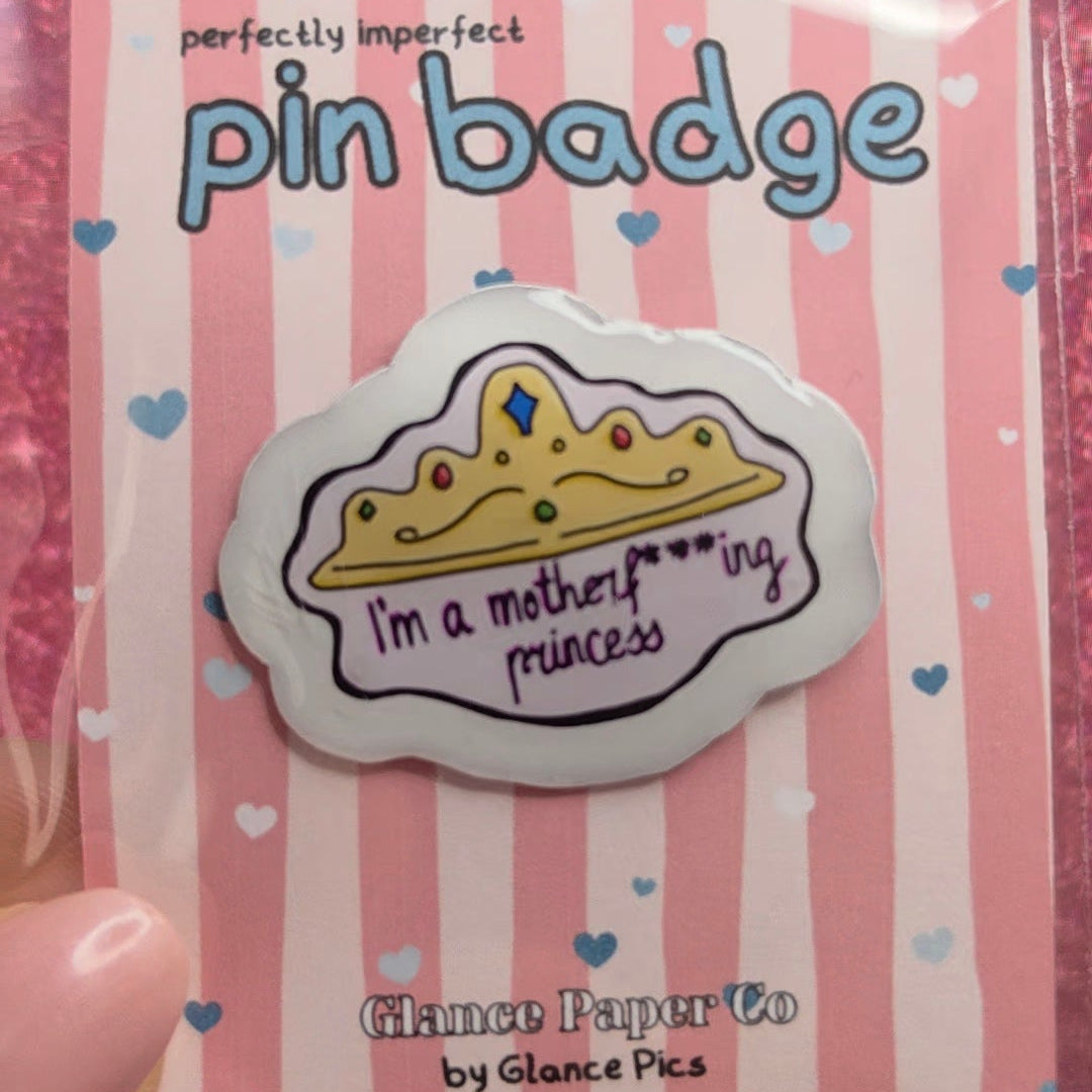 A hand holding a pink and white striped card displaying a pin badge. The badge features an illustration with a gold crown and the text 'I'm a motherly princess'.