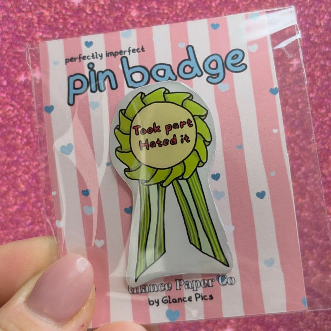 A hand with pink manicured nails holds a handmade resin pin badge. The badge features a green ribbon award design with a yellow center that reads 'Took part, hated it"