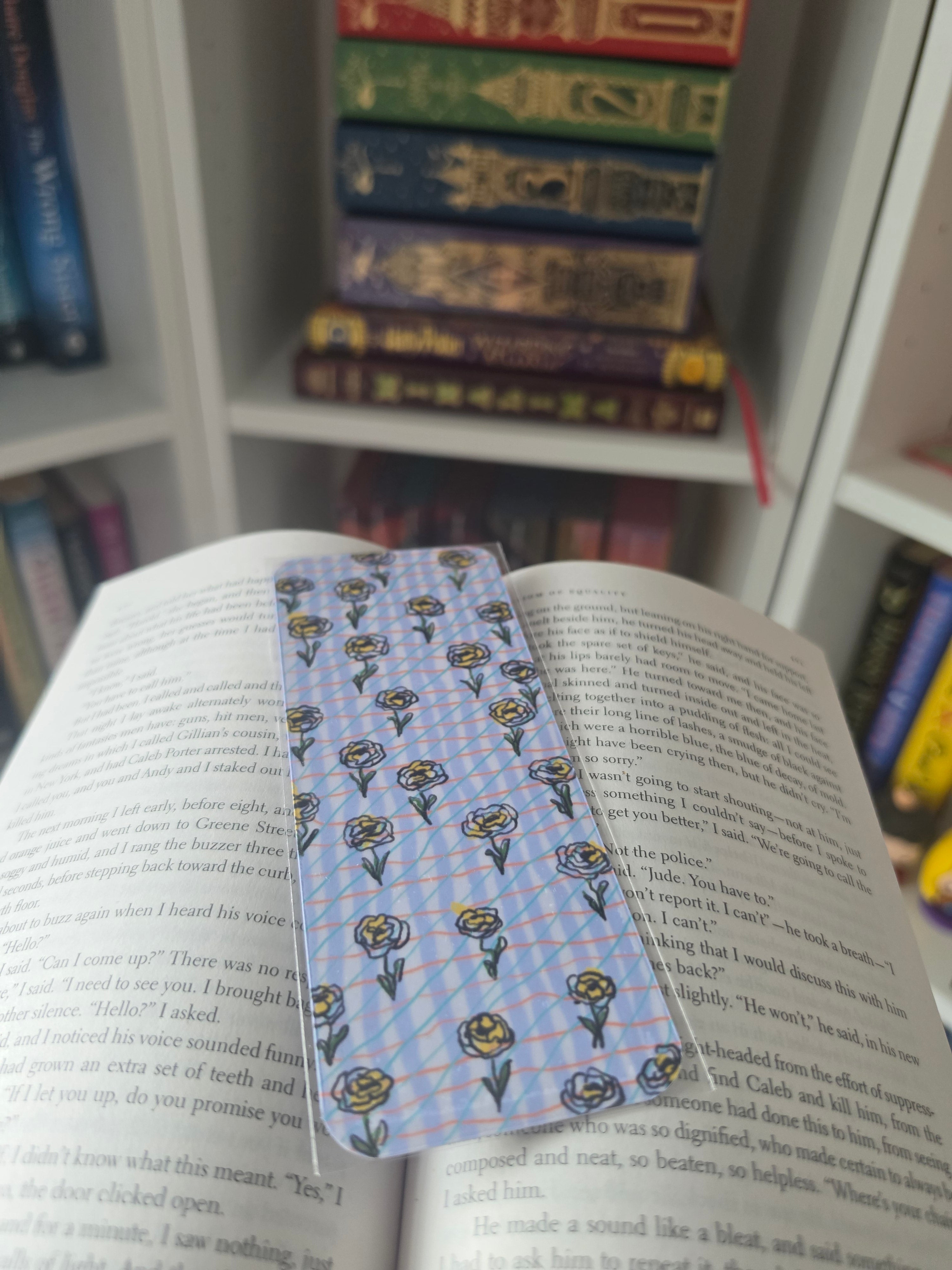 A light blue patterned bookmark with yellow and green floral designs is placed across open book pages, with a stack of books visible in the blurred background.