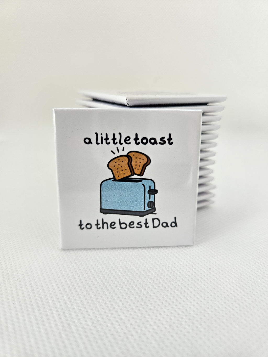 magnet with a design of a toaster and toast, text 'to the best Dad' and 'a little toast', on a white textured background.