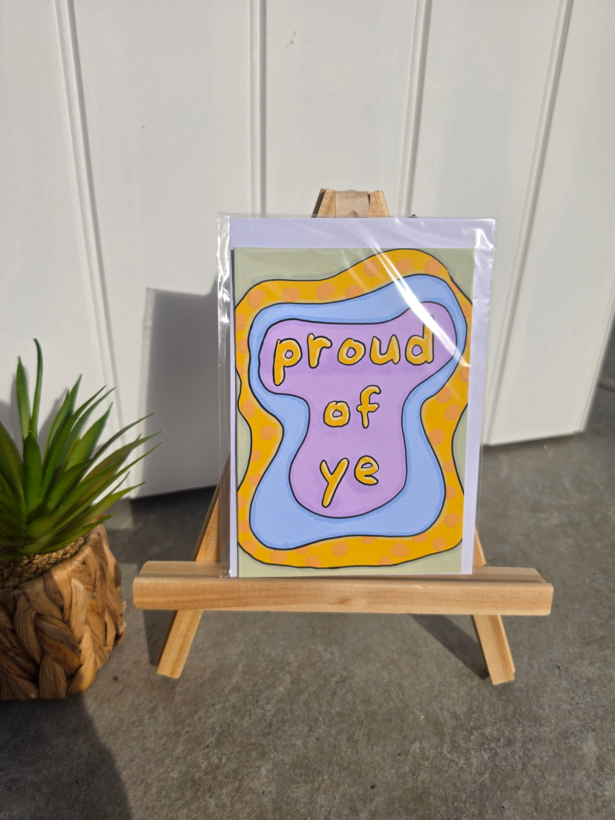 A greeting card with a retro wavy design reading 'proud of ye' in yellow text, displayed on a small wooden easel. The card features a purple center, light blue waves, a mustard yellow border, and a sage green background.