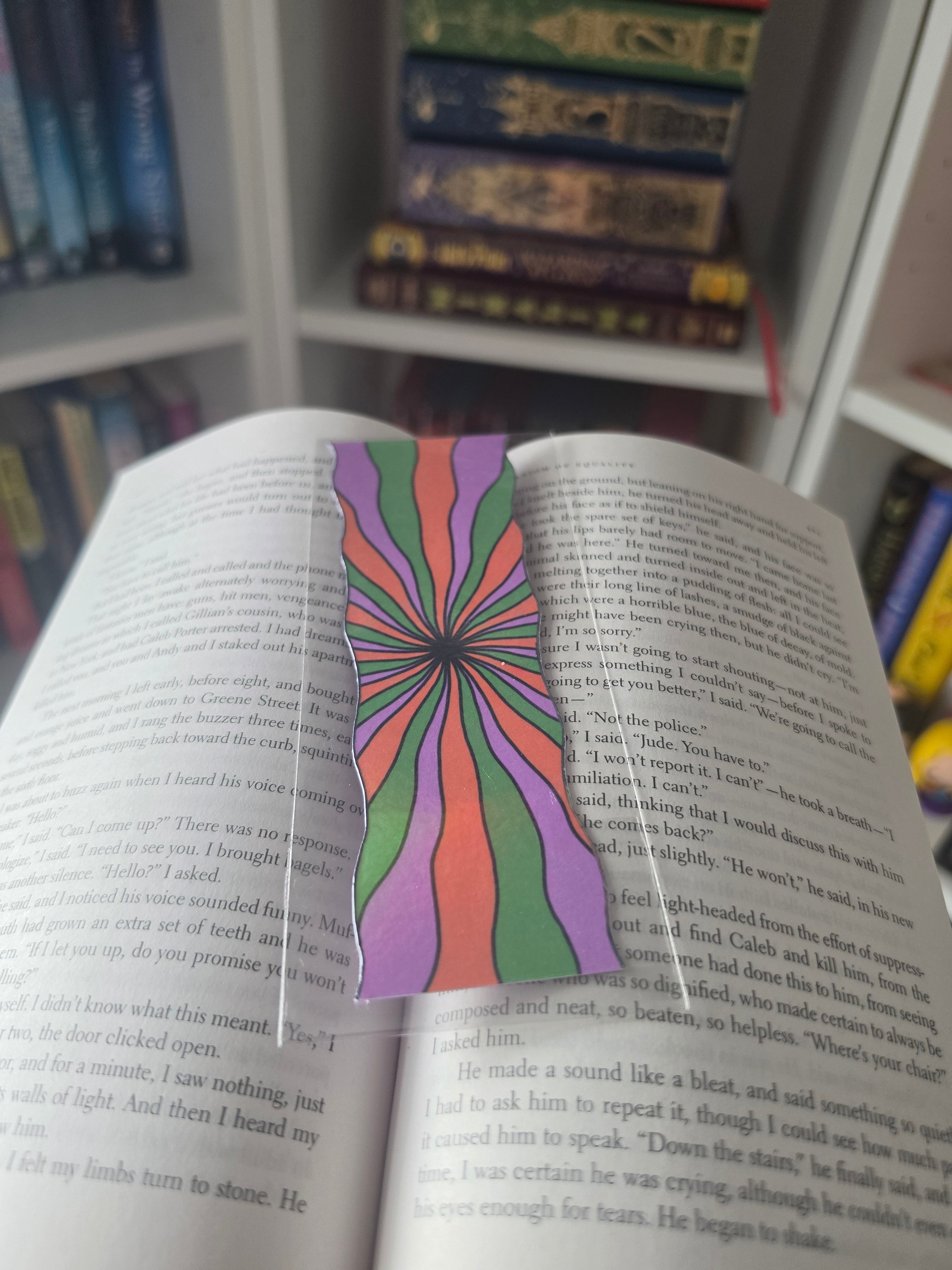 A colorful spiral-patterned paper bookmark with a psychedelic rainbow design lies on an open book, with other books blurred in the background.