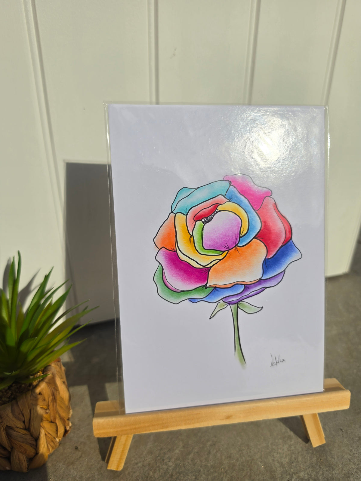 A square art print featuring a colorful rainbow peony, displayed on a small wooden easel. The print is wrapped in a protective sleeve, set against a background of white paneled walls and a gray floor, with a small potted plant on the left
