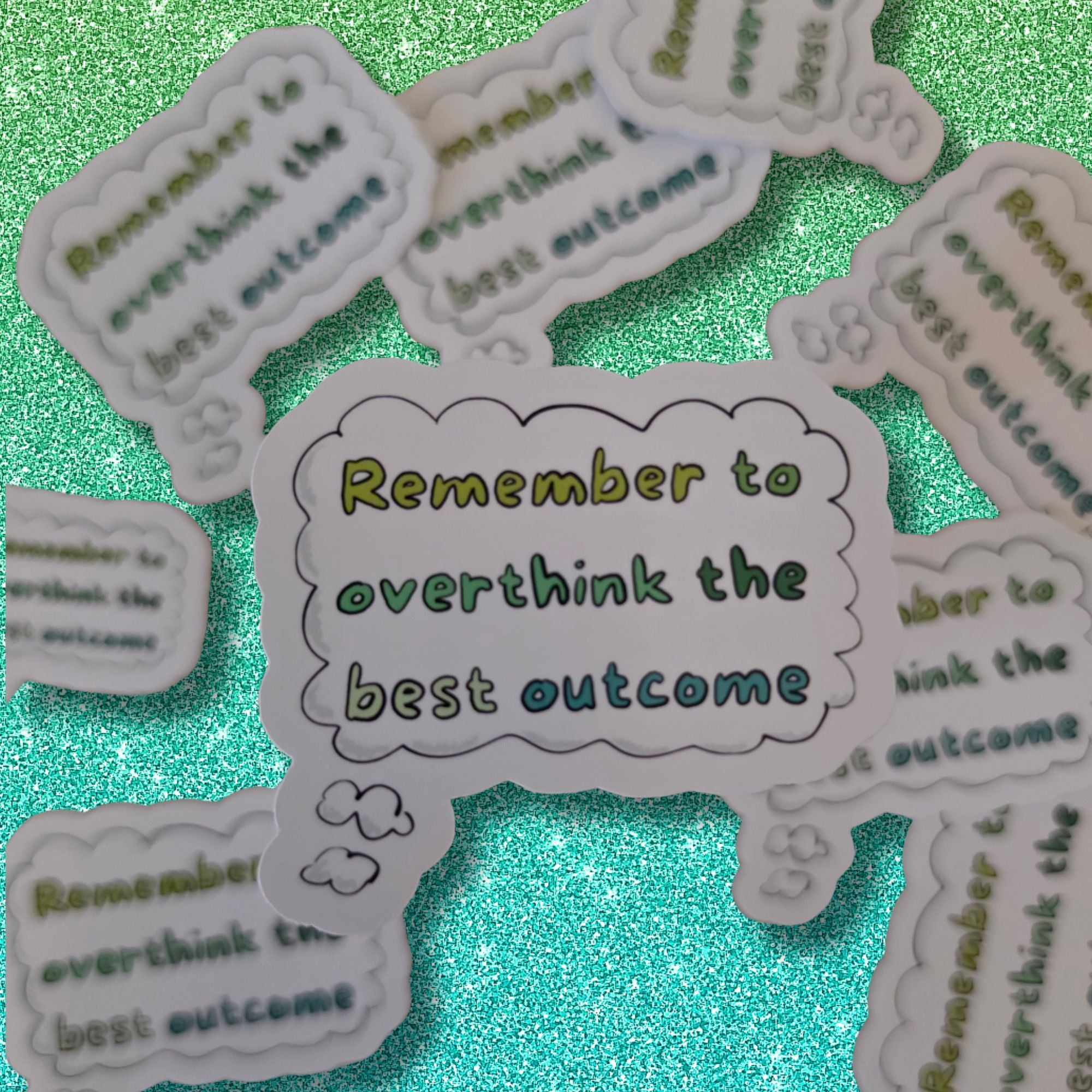 A collection of die-cut cloud-shaped stickers with the motivational phrase 'Remember to overthink the best outcome' in green and yellow lettering, arranged on a sparkling teal background.