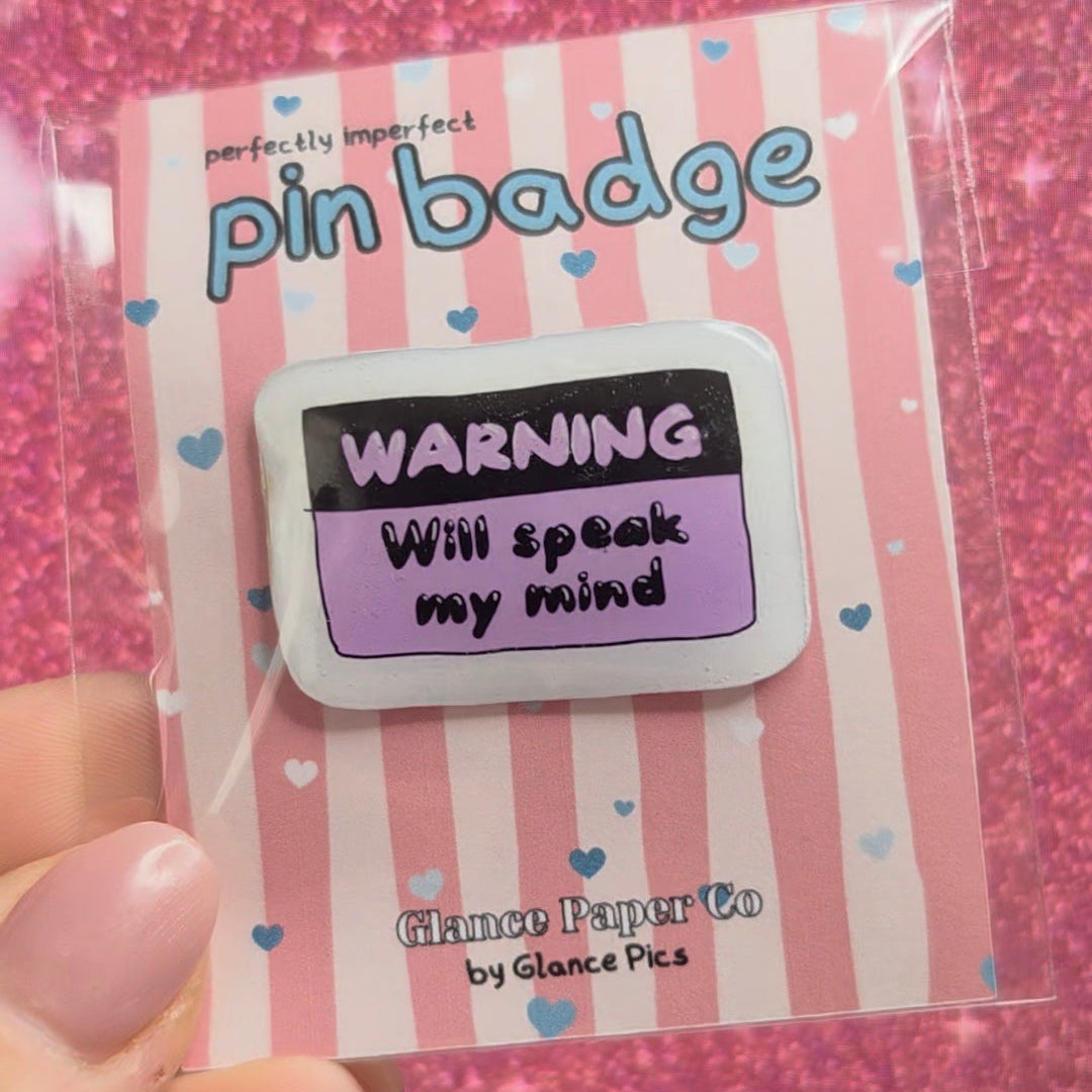 A hand holding a pink and white striped pin badge card featuring a warning-style pin that reads 'WARNING Will speak may mind' on a glittery pink background.