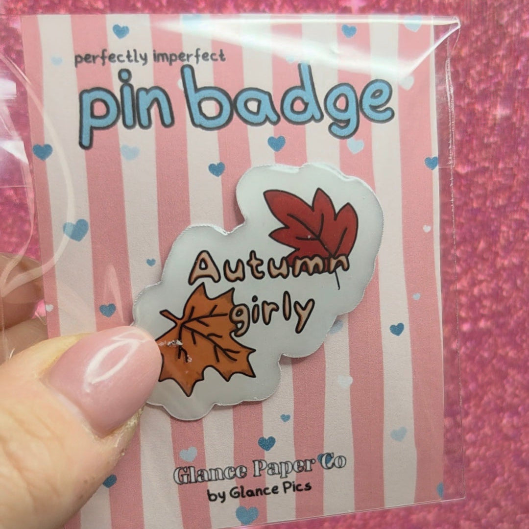 A hand with pink manicured nails holds a packaged pin badge. The badge is shaped like autumn leaves and says 'Autumn girly' with orange and brown leaves on a white background.