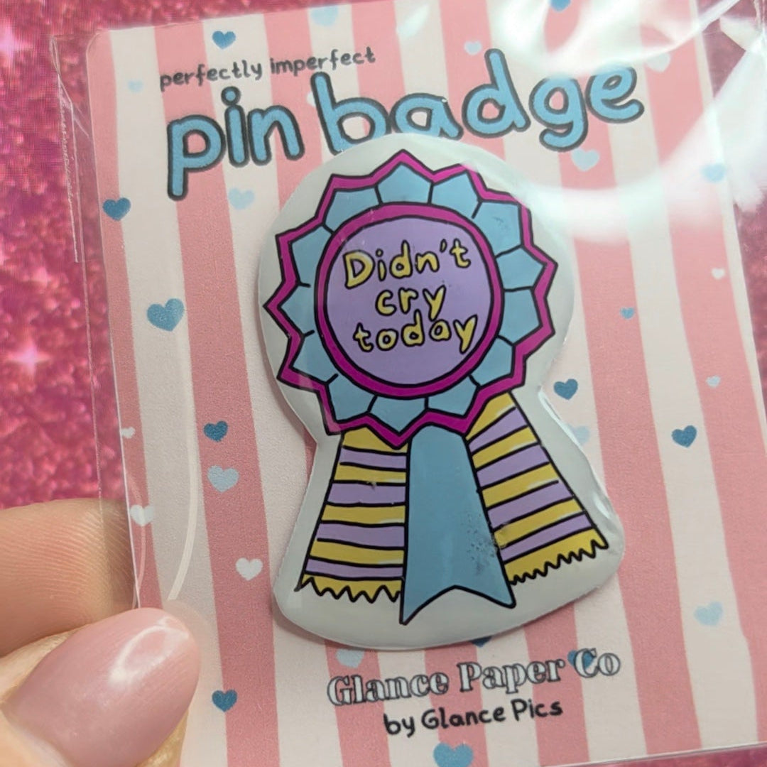 A hand holding a packaged resin pin badge on a pink glittery background. The pin badge is a ribbon award design with the text 'Didn't cry today' in yellow on a purple center, with pink, blue, and yellow details.