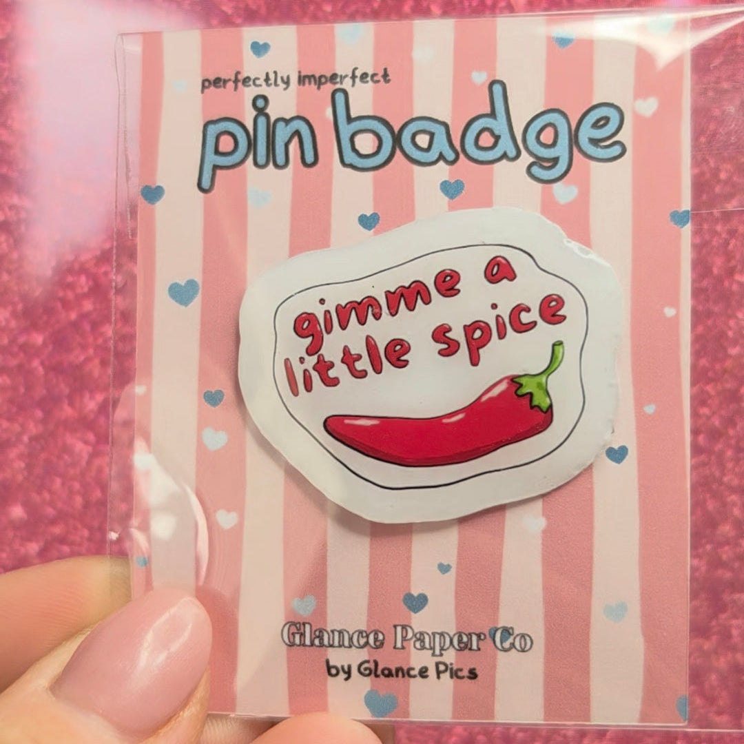 A hand holds a clear plastic package containing a pin badge. The badge features a white background with a pink and white striped border, the text 'pin badge' in blue, and a central white shape with the phrase 'gimme a little spice' in red script around a red chili pepper illustration.