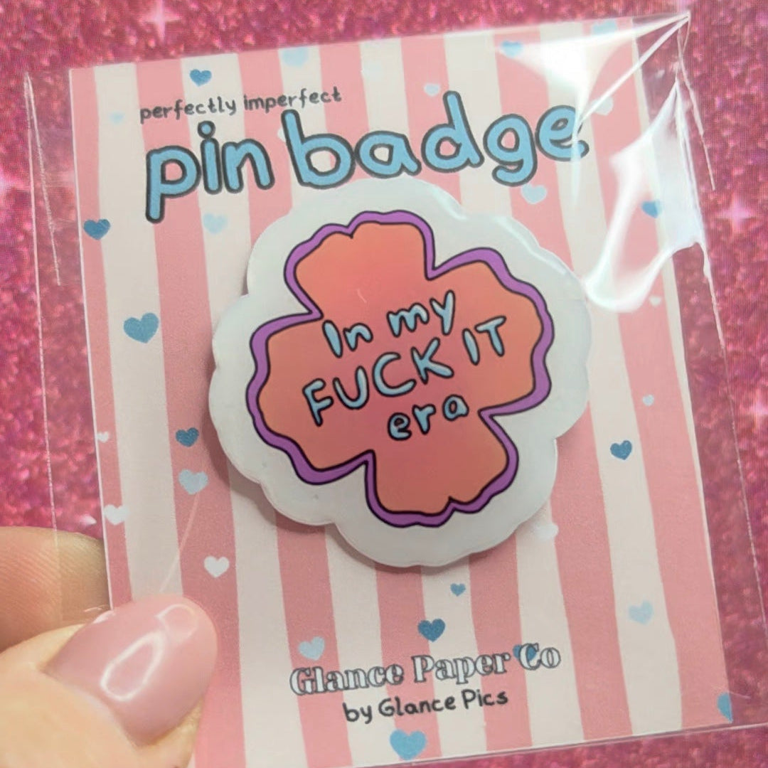 A hand holding a pink and white striped card displaying a resin pin badge that reads 'In my FUCK IT era'. The card has 'perfectly vegan-feel pin badge' at the top and 'Glance Paper Co by Glance Pics' at the bottom.