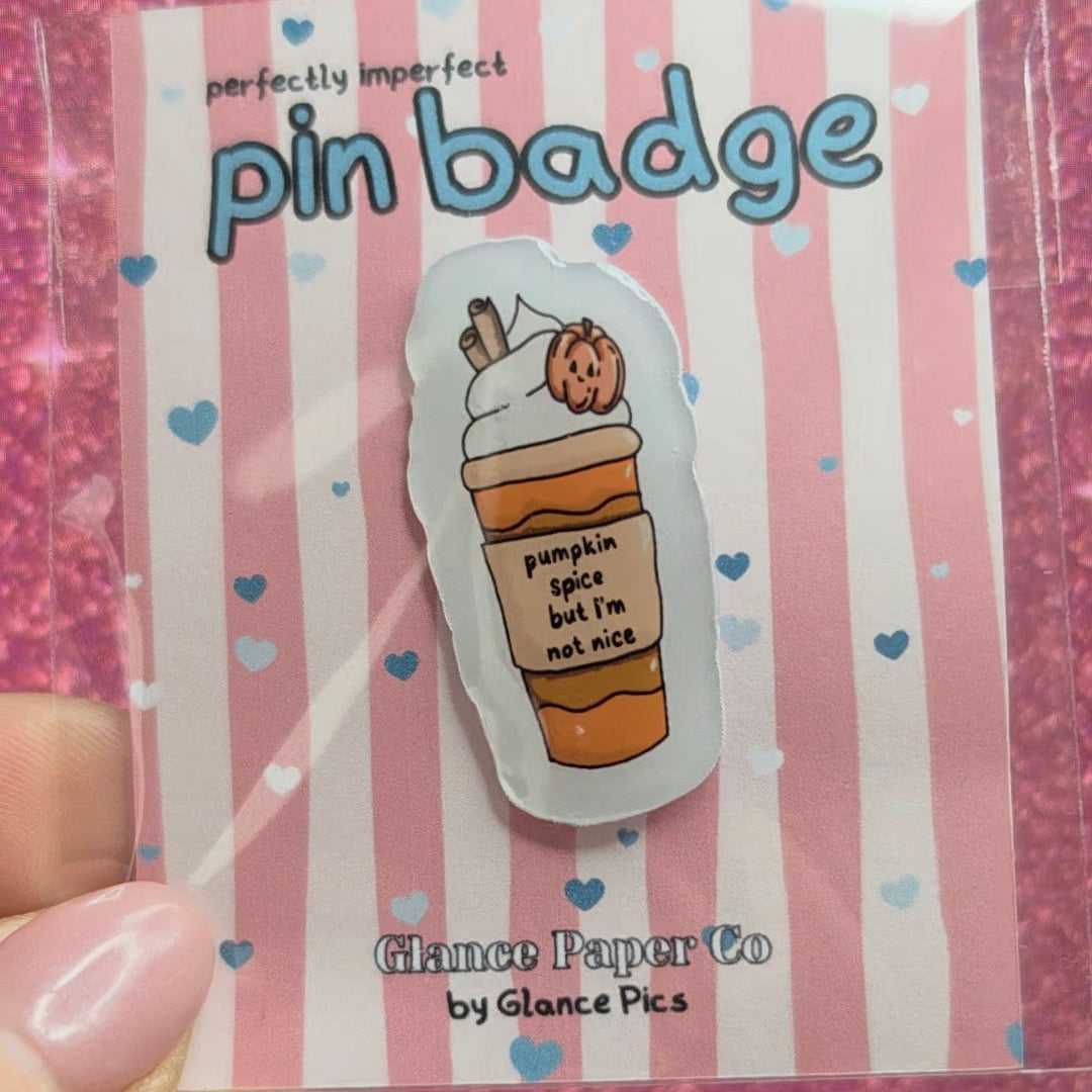 hand holding a handmade resin pin badge, on a pink striped display card. The pin is shiny with an images of a handdrawn pumpkin spiced latte which states "pumpkin spice but I'm not nice" 