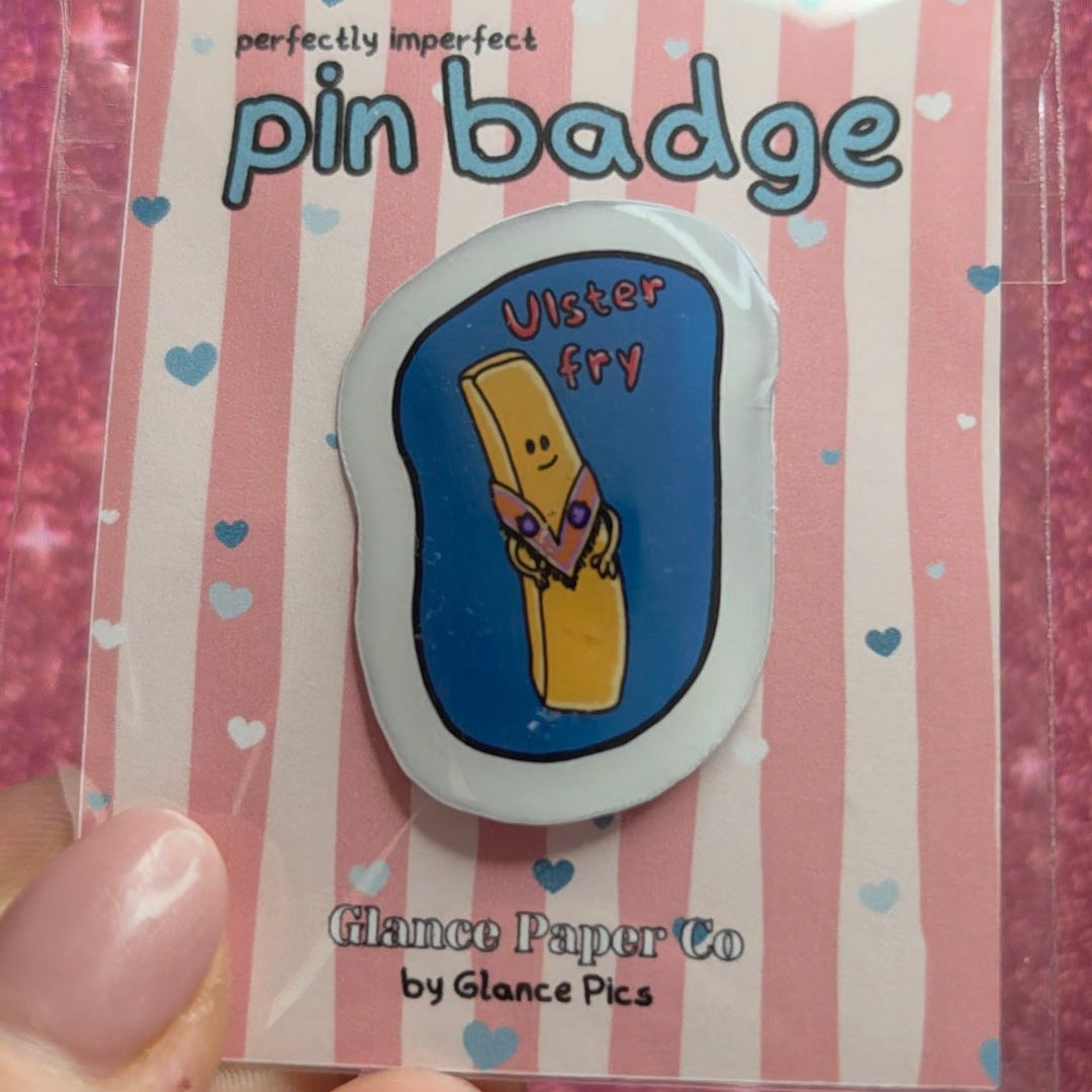 A hand holding a packaged enamel pin badge on a pink glittery background. The pin badge features a yellow french fry wearing an orange order sash with inside a blue oval, mounted on a pink and white striped backing card labeled 'pin badge' and 'Glance Paper Co by Glance pics