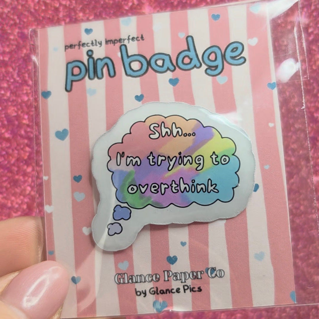 A hand holding a packaged pin badge from Glance Paper Co by Glance Pics. The resin pin badge is a pastel rainbow-colored speech bubble with the text 'Shh...i'm trying to overthink"