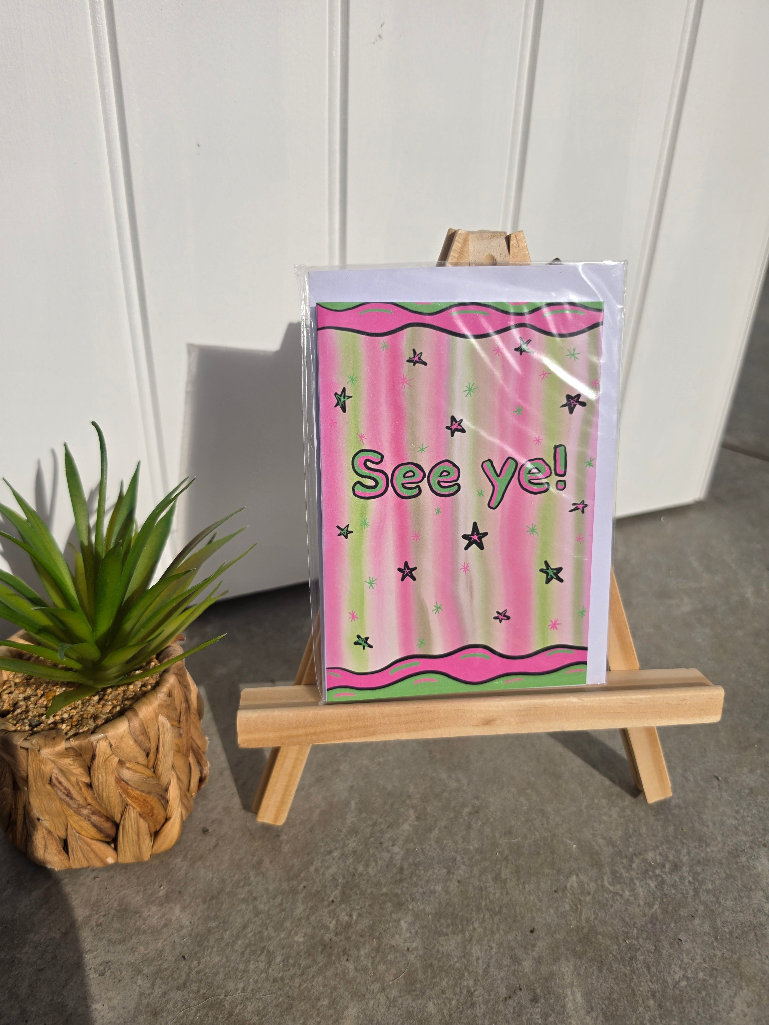 A small greeting card with a pink and green striped design, black stars, and the text 'See ye!' displayed on a miniature wooden easel. A potted succulent sits to the left, with a white paneled wall and concrete floor in the background.