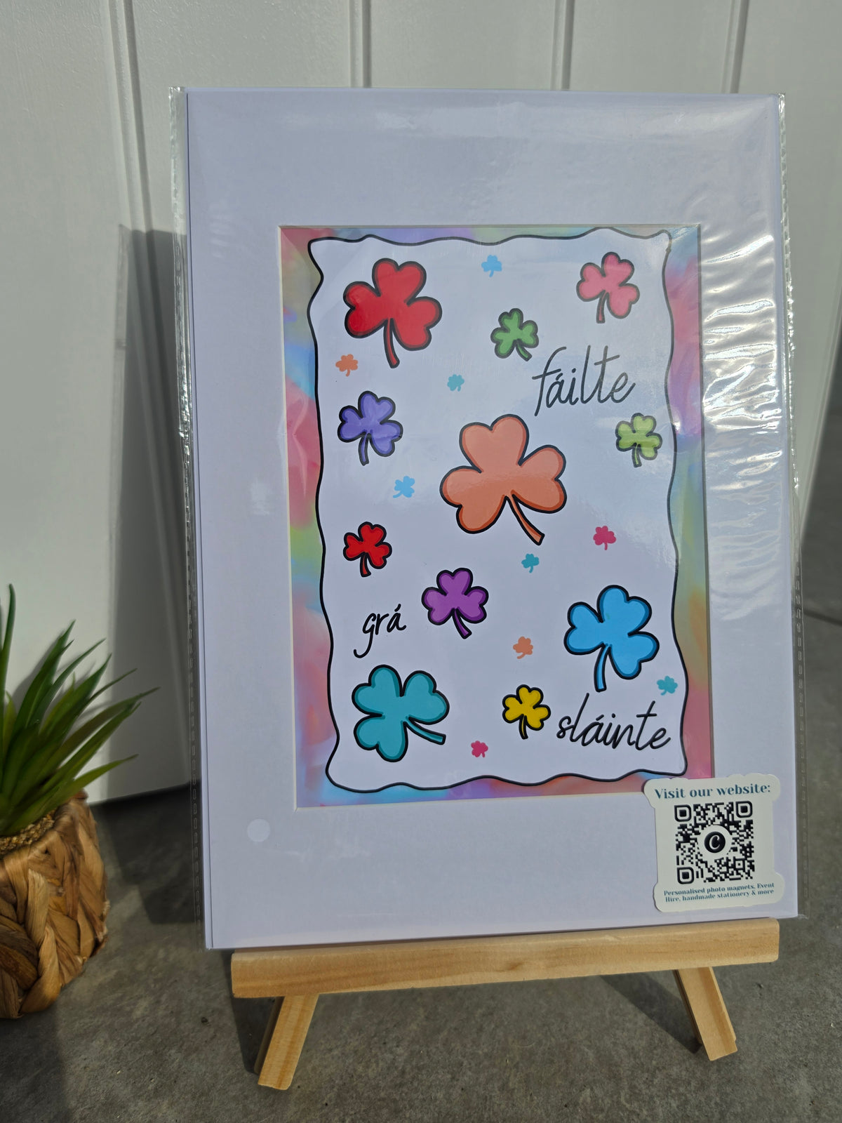 A square printed art piece or card with colorful shamrock illustrations and the words 'falte grä slånte' on a white sheet, photographed standing on a small wooden easel against a light paneled wall. A small QR code sticker is visible in the lower right corner.