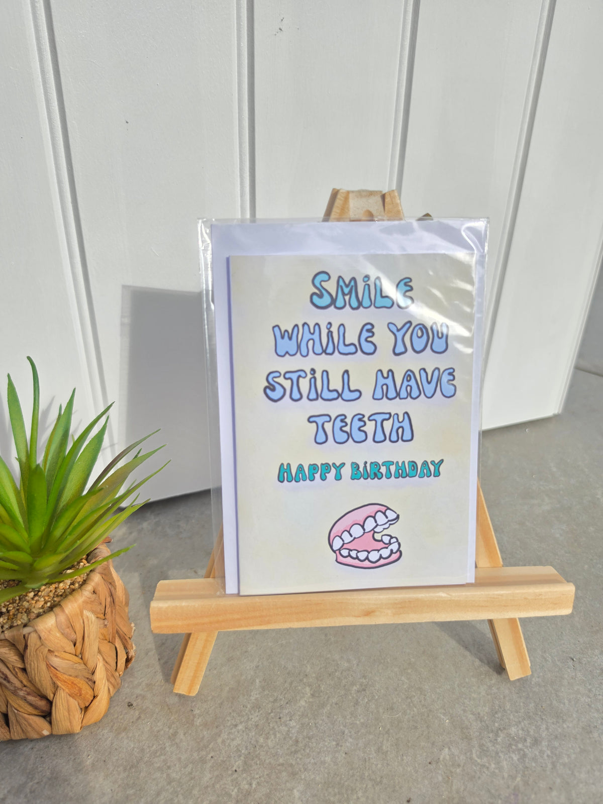 A square birthday card with the text 'SMILE WHILE YOU STILL HAVE TEETH HAPPY BIRTHDAY' and a chomp graphic, displayed on a small wooden easel. The card is wrapped in clear plastic, set against a white paneled wall with a small potted succulent on the left.