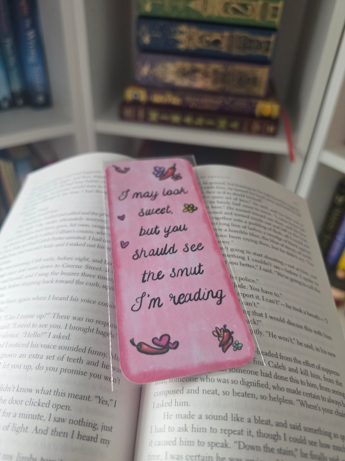 A pink rectangular book marker with a clear plastic sleeve and printed quote reading 'I may look sweet, but you should see the smut I'm reading' with small butterfly and heart illustrations, placed on an open book with other books blurred in the background.