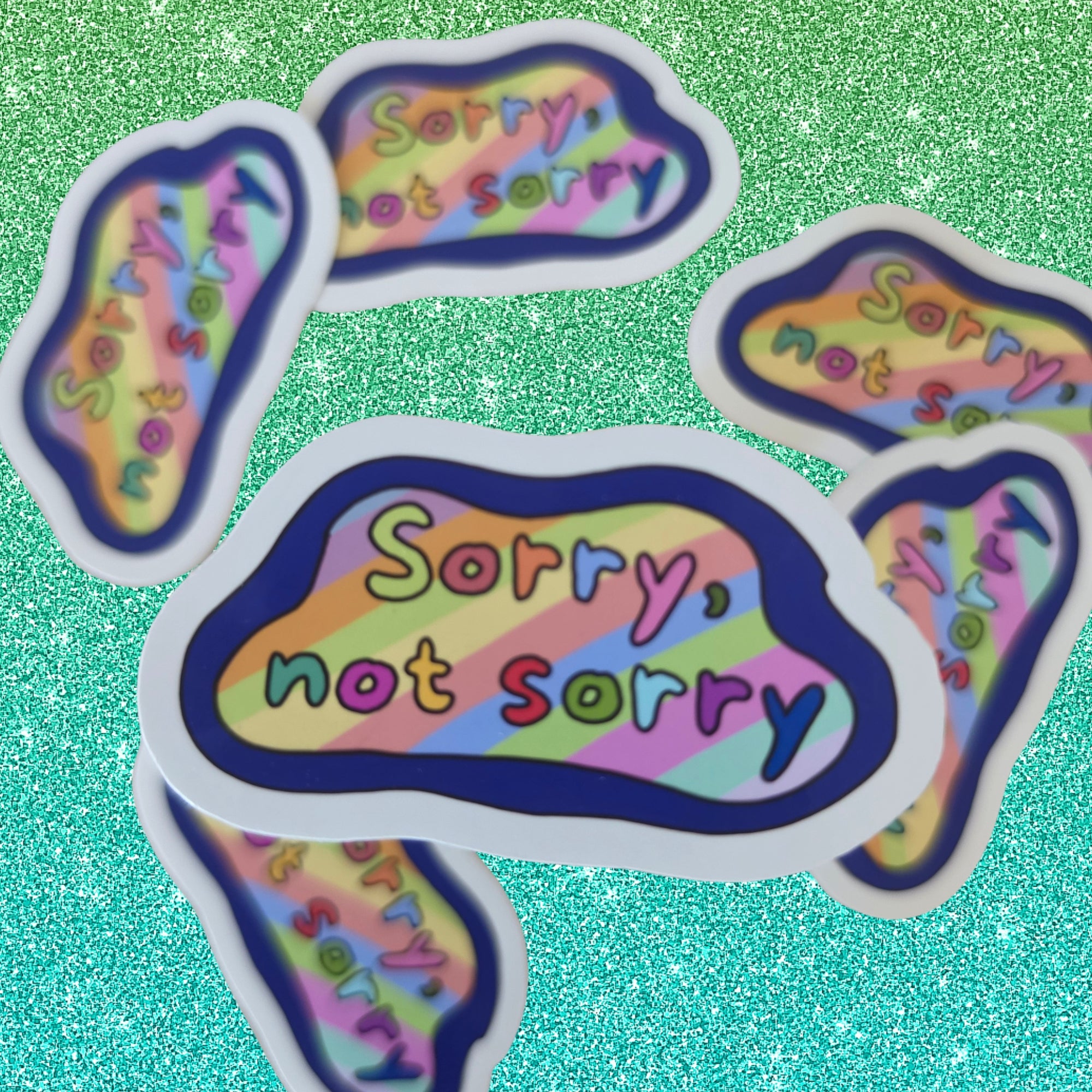 Five cloud-shaped stickers with the text 'Sorry, not sorry' in colorful bubble letters on a pastel rainbow striped background, arranged on a glittery green surface.