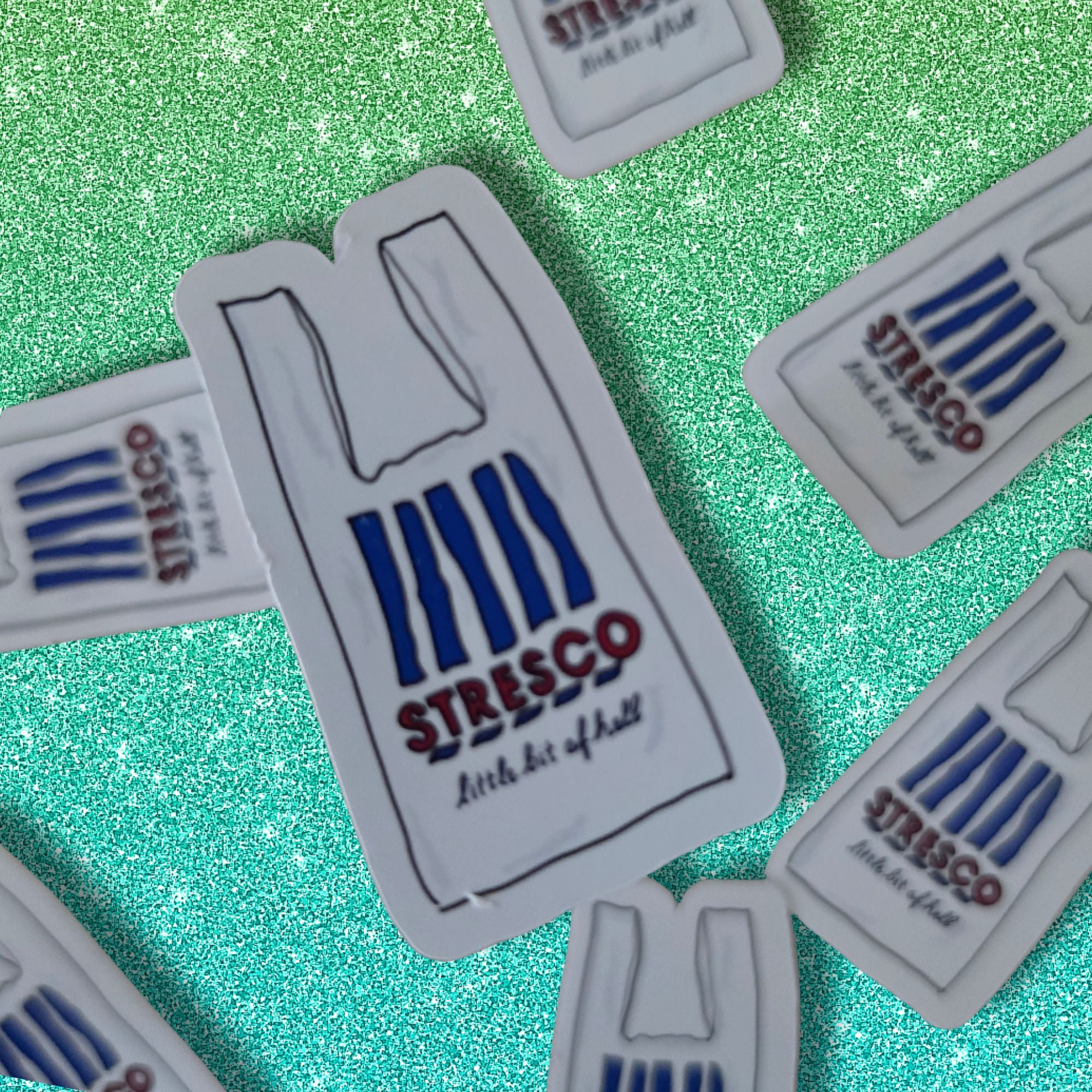 Several small white stickers shaped like a Stresco bag are scattered on a glittery green background. The central sticker is fully visible and shows the Stresco logo in blue and red with the phrase 'little bit of hell'.