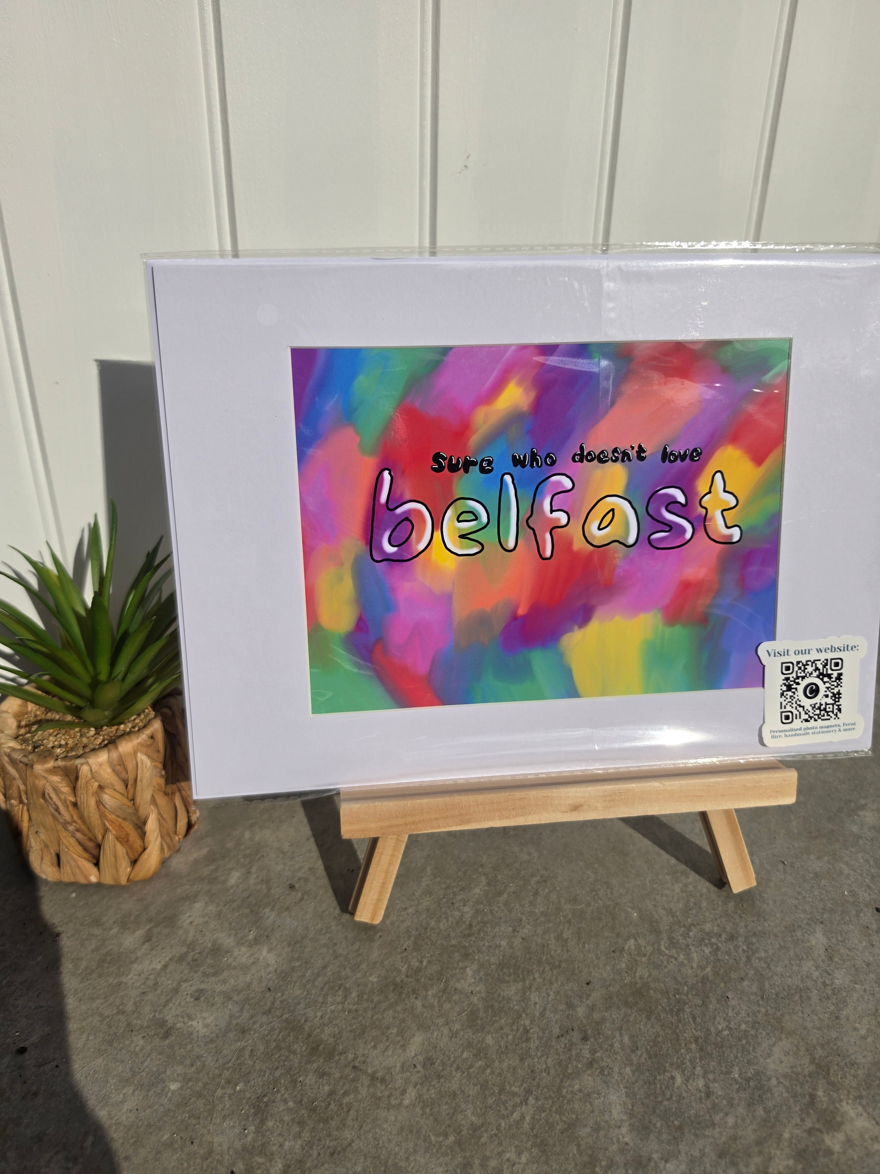 A framed art print displayed on a small wooden easel. The print features a colorful abstract background with the text 'Sure who doesn't love belfast' in stylized lettering.