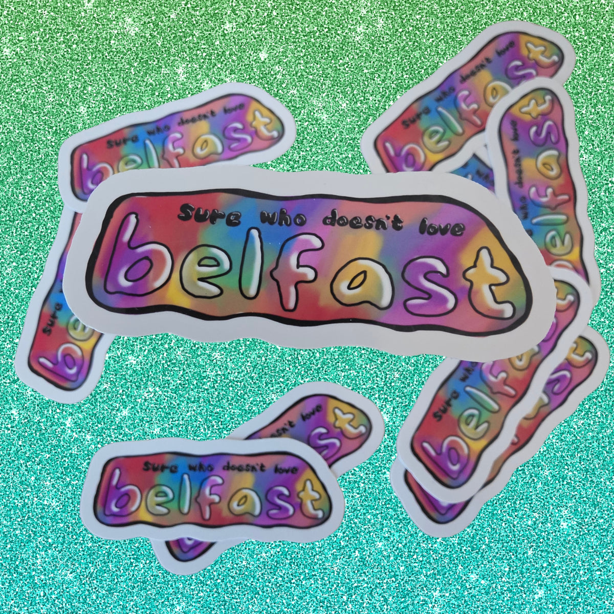 A set of die-cut stickers featuring the phrase 'sure who doesn't love belfast' in white bubble letters over a rainbow gradient background, arranged on a sparkling teal surface.