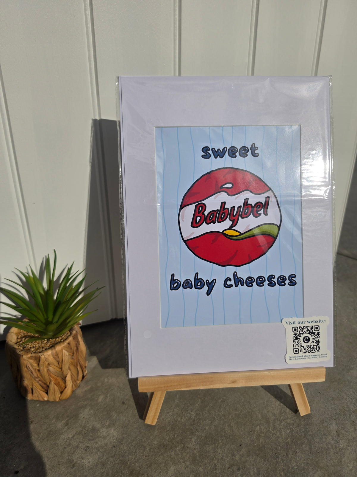 A framed novelty sign or print displayed on a small wooden easel, featuring the text 'sweet baby cheeses' around a red and white Babybel logo illustration. The sign is wrapped in protective plastic, set against a paneled wall with a small potted plant to the left.