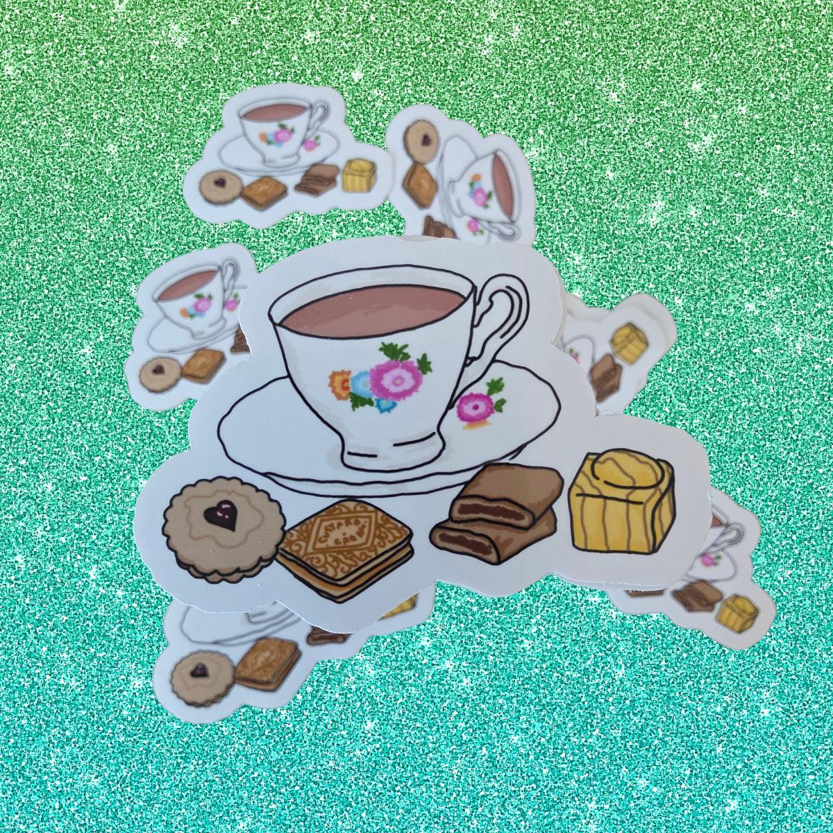 A die-cut sticker featuring a floral teacup and saucer surrounded by assorted pastries and chocolates, displayed on a sparkling green glitter background.