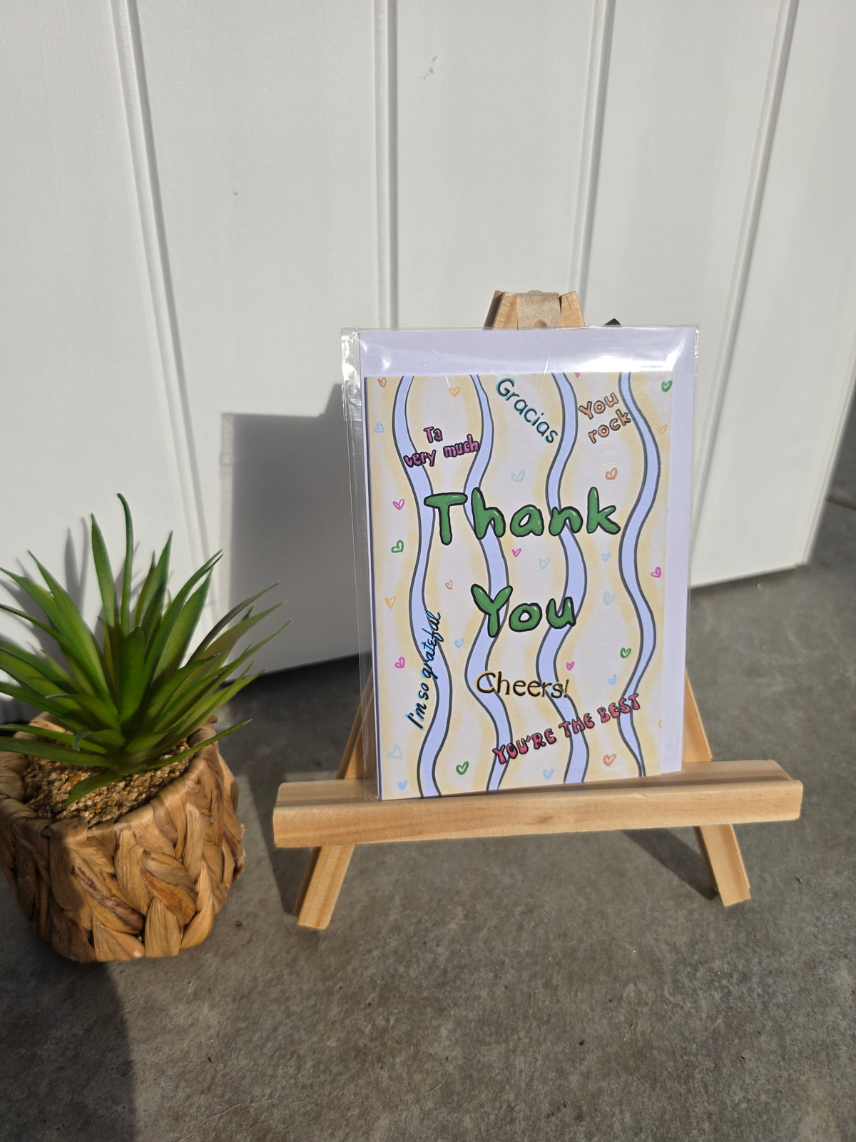A greeting card displayed on a small wooden easel, next to a potted green plant. The card features a white background with wavy blue and yellow lines and colorful text reading 'Thank You', 'To my family', 'Gracias', and 'Cheers!'.