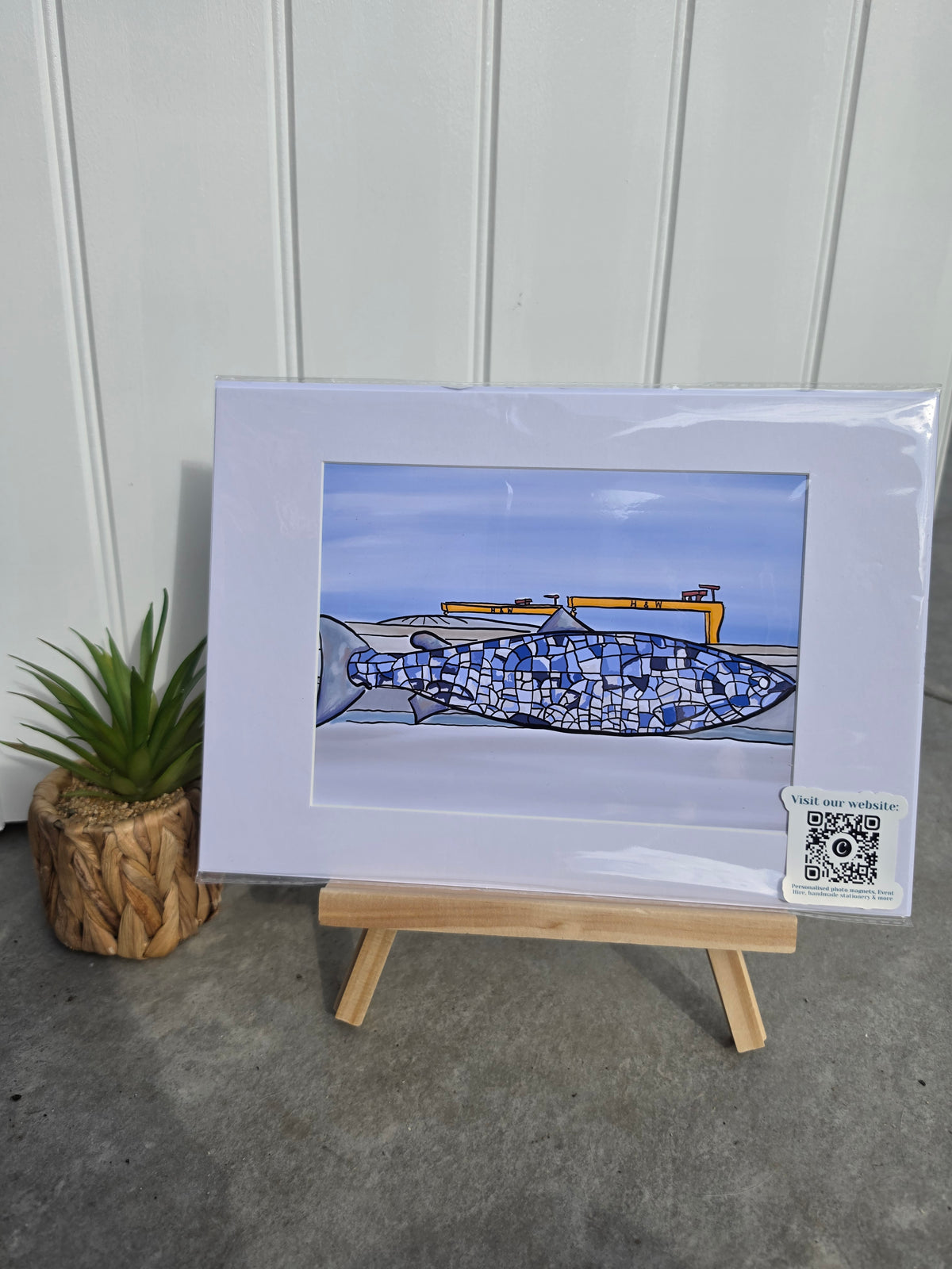 A framed art print of a blue and white fish with a yellow boat in the background, displayed on a small wooden easel against a white paneled wall with a small potted plant to the left.