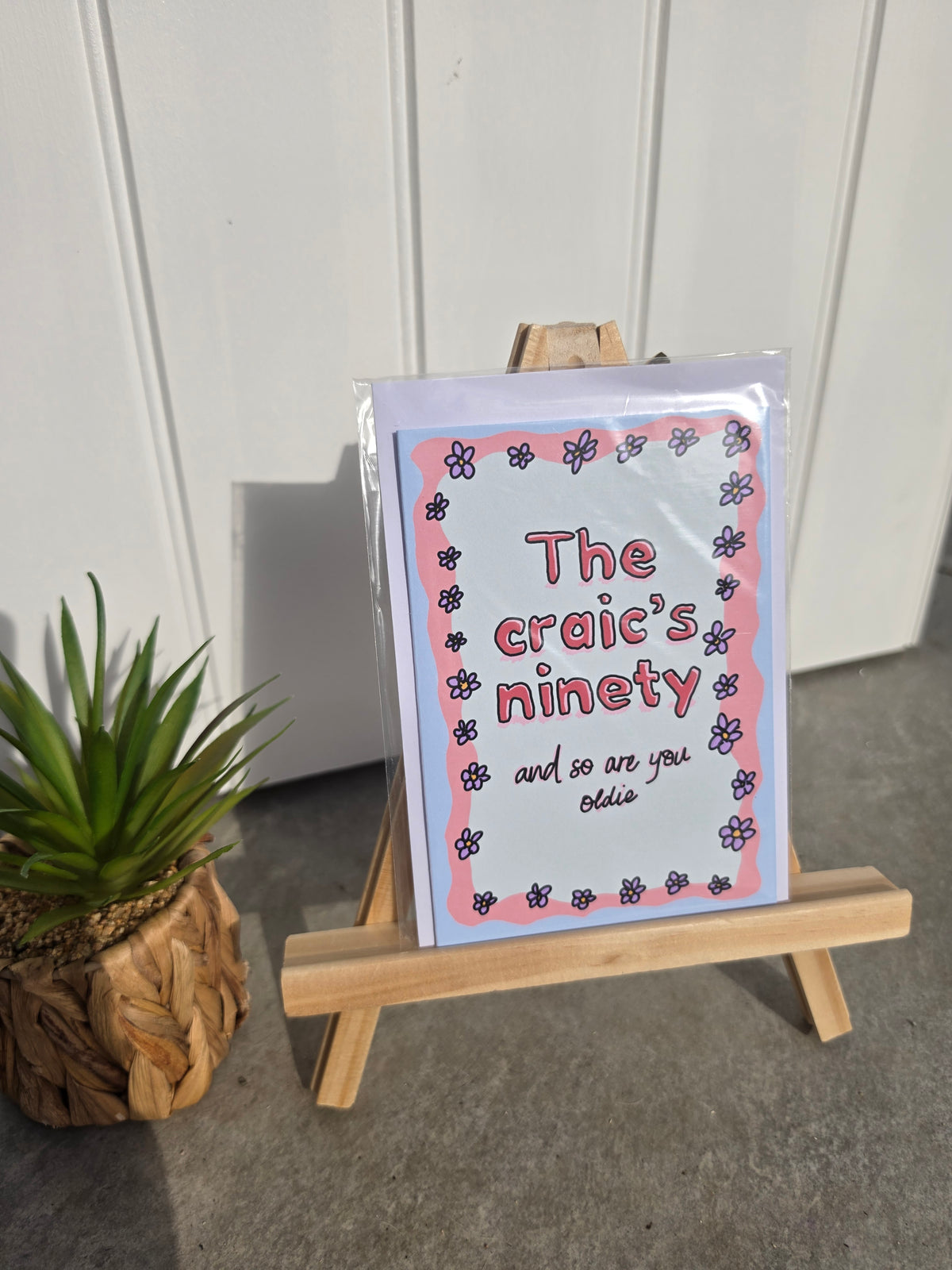 A greeting card displayed on a small wooden easel, with a potted plant to the left and a white paneled wall in the background. The card reads 'The craic's ninety and so are you oldie' with a pink and blue floral border.