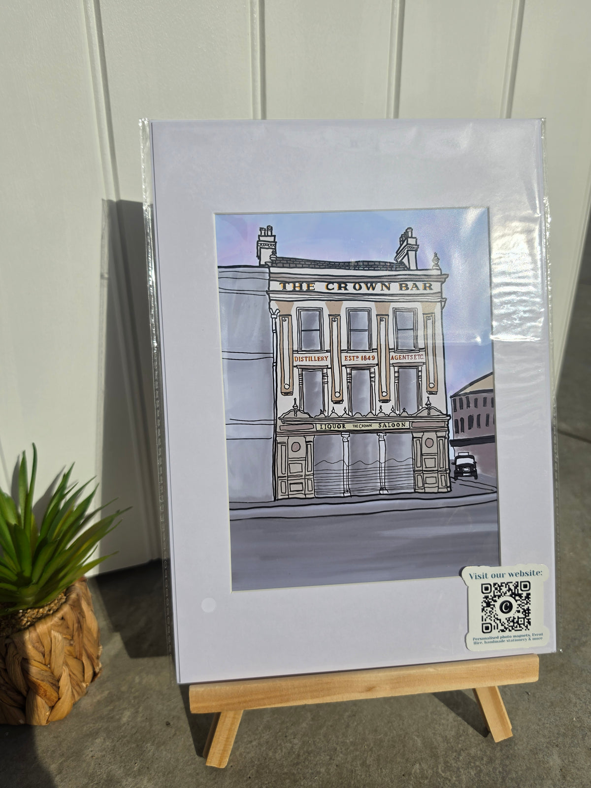 A matted art print of a hand-drawn illustration of 'The Crown Bar' pub, displayed on a small wooden easel next to a potted plant.