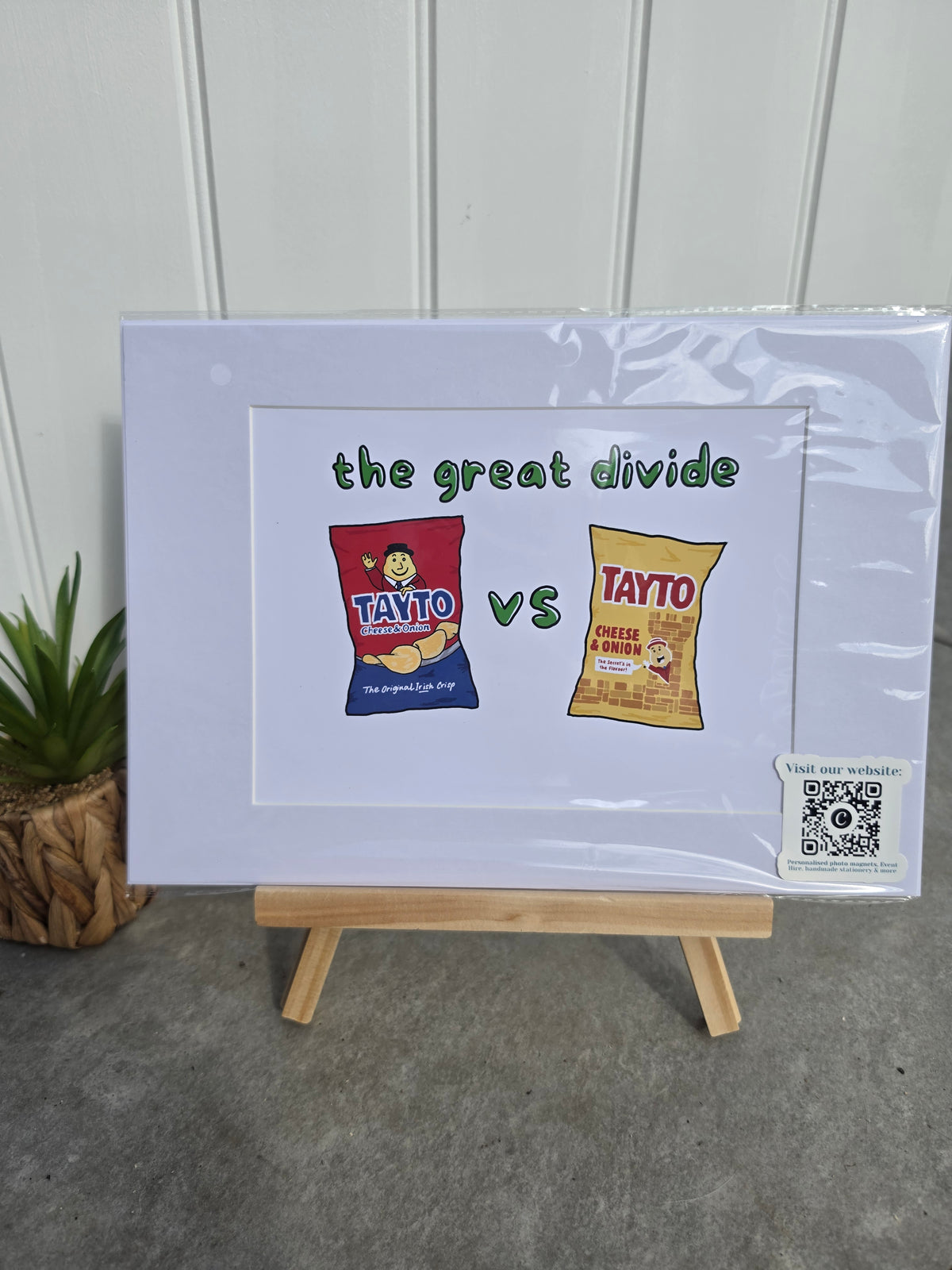 A framed print on a small wooden easel, featuring a comparison graphic titled 'the great divide' with a red Tayto chip bag versus a yellow Tayto chip bag 