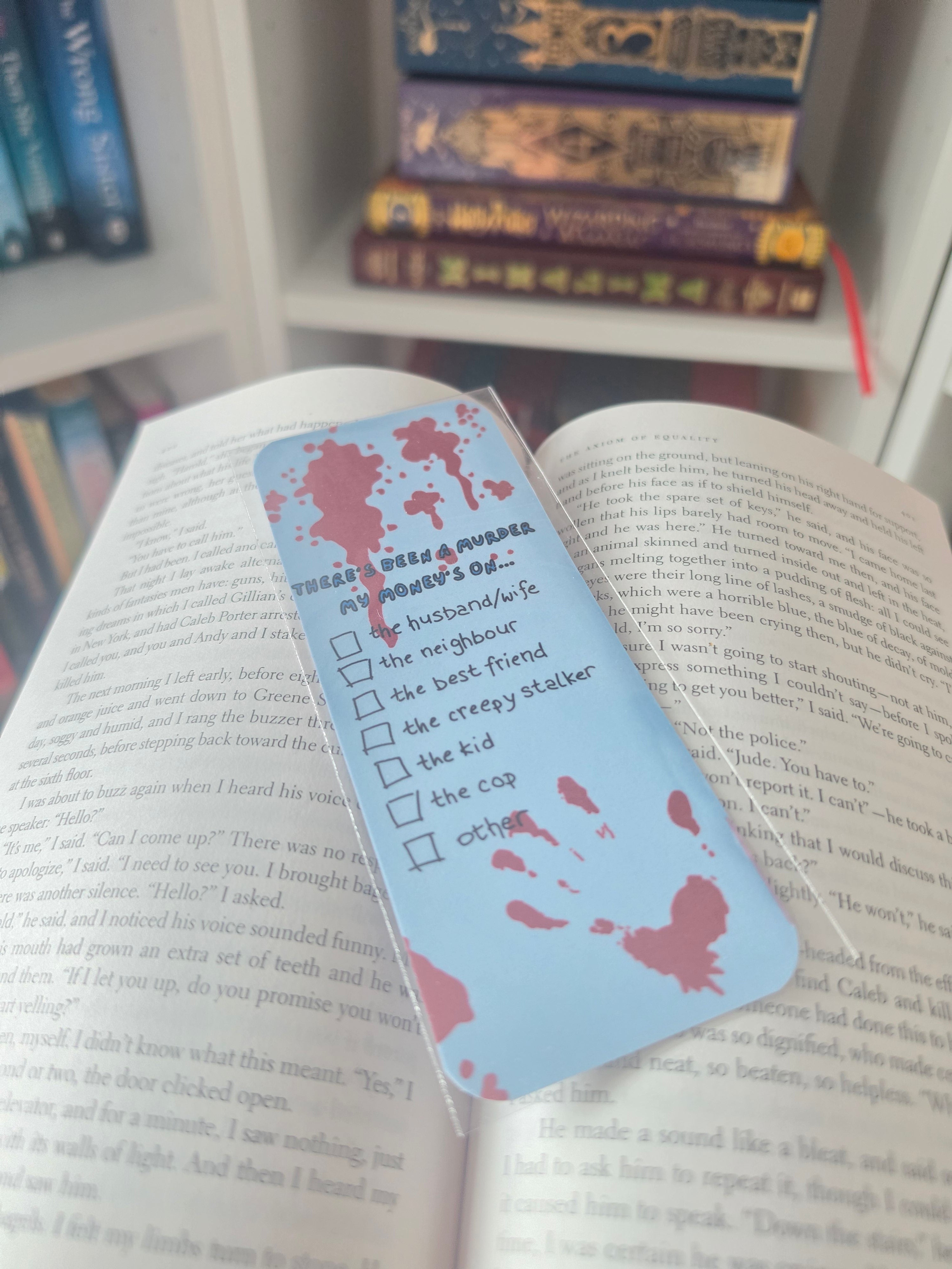 A light blue checklist bookmark with red blood splatter details, reading 'there's been  a murder' and listing checkboxes like 'the husband/wife', 'the neighbor', 'the best friend', 'the creepy statistic', 'the kid', 'the cap', and 'other'. It is shown resting on an open book with other books blurred in the background.