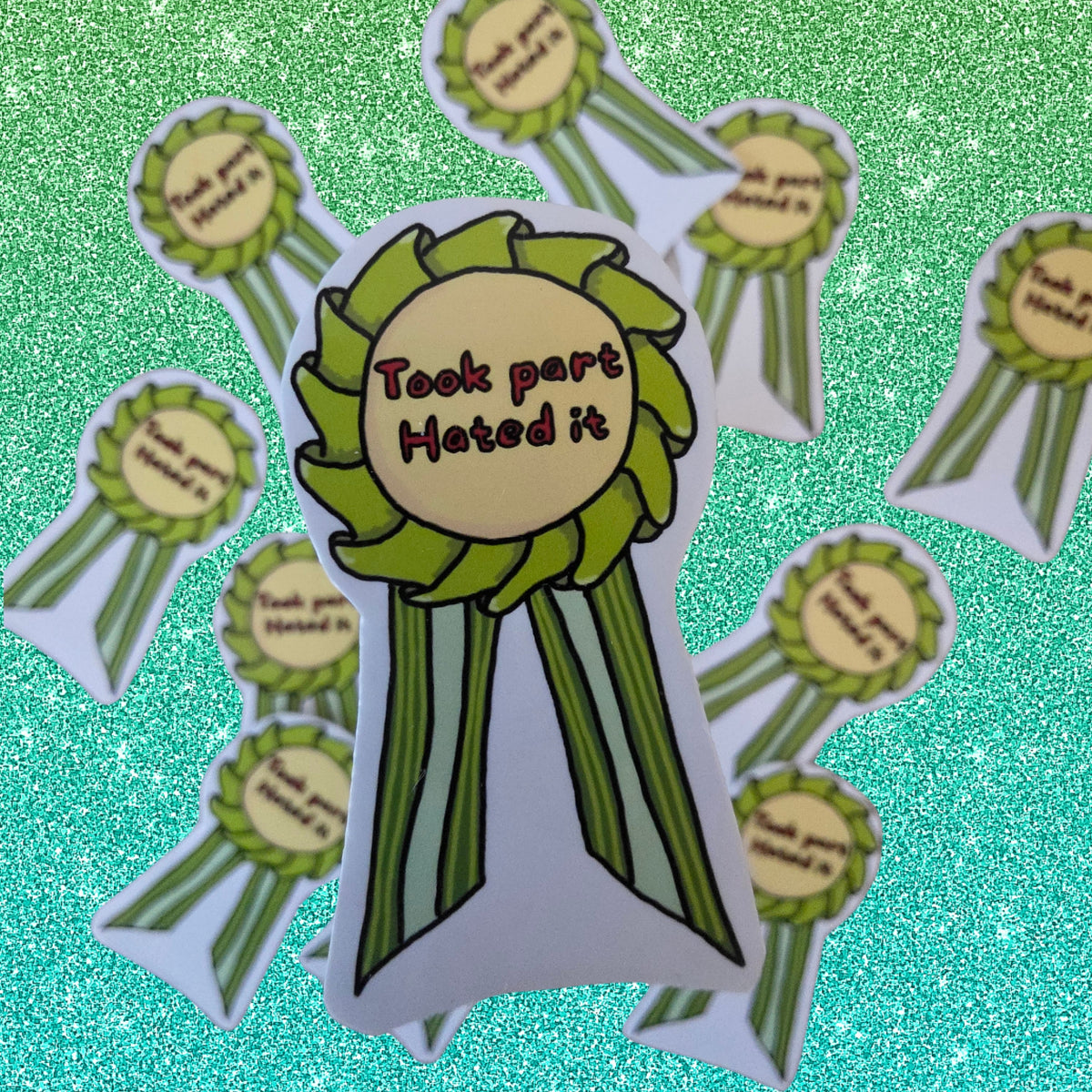 A die-cut sticker shaped like a green and white rosette ribbon with the text 'Took part Hated it' in the center, surrounded by several identical rosette stickers on a glittery turquoise background.