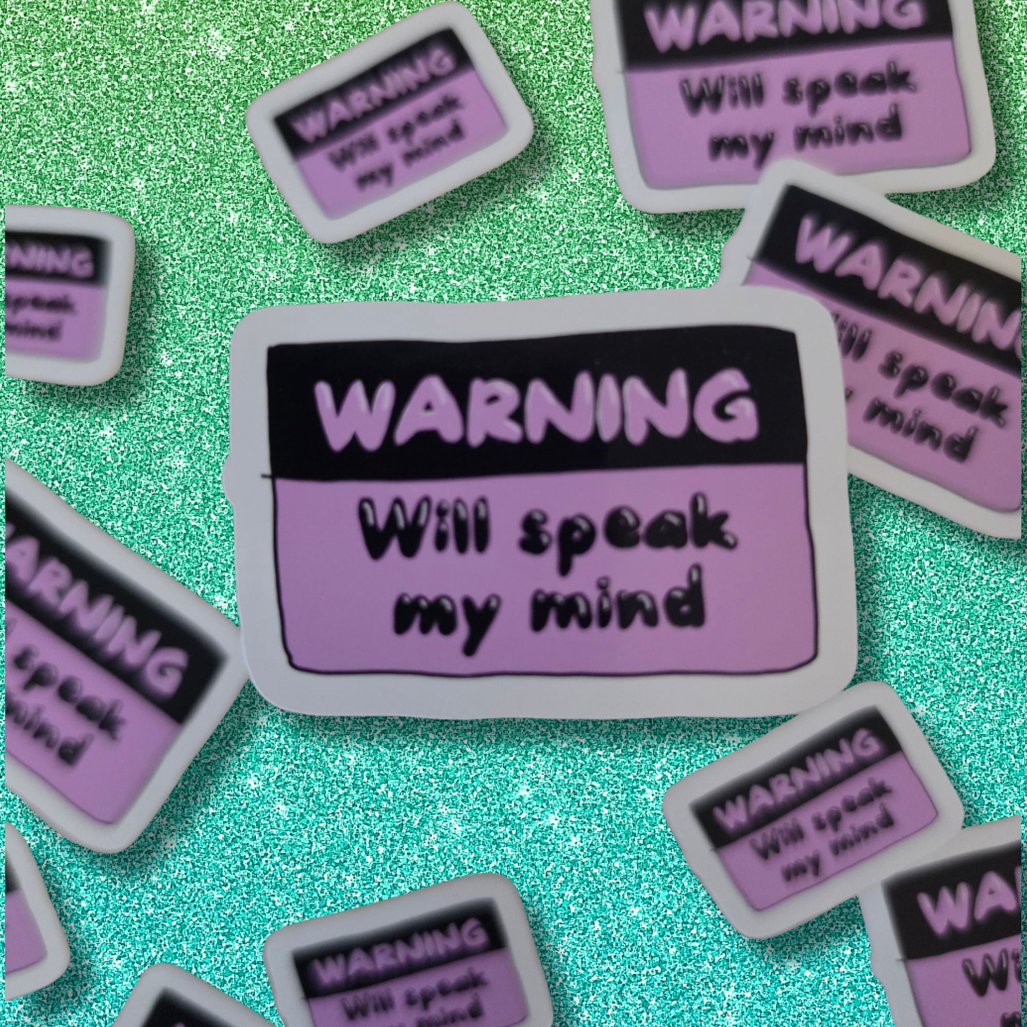 A close-up of multiple matching warning label stickers scattered on a glittery teal background. The central sticker reads “WARNING Will speak my mind” in black text on a pink and black label with a white border.
