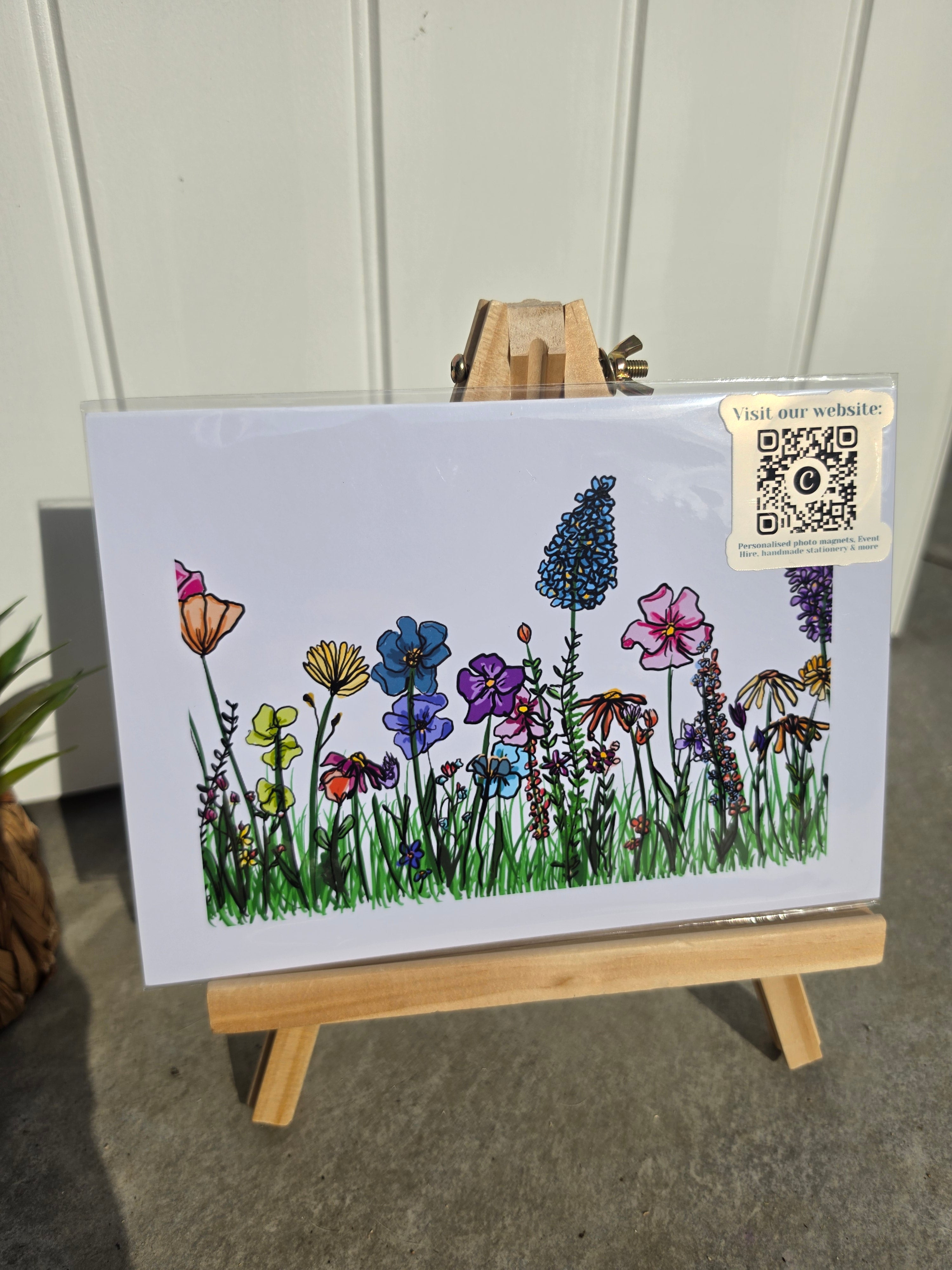 An art print featuring a colorful wildflower meadow illustration, displayed on a small wooden easel. The print has a white border 