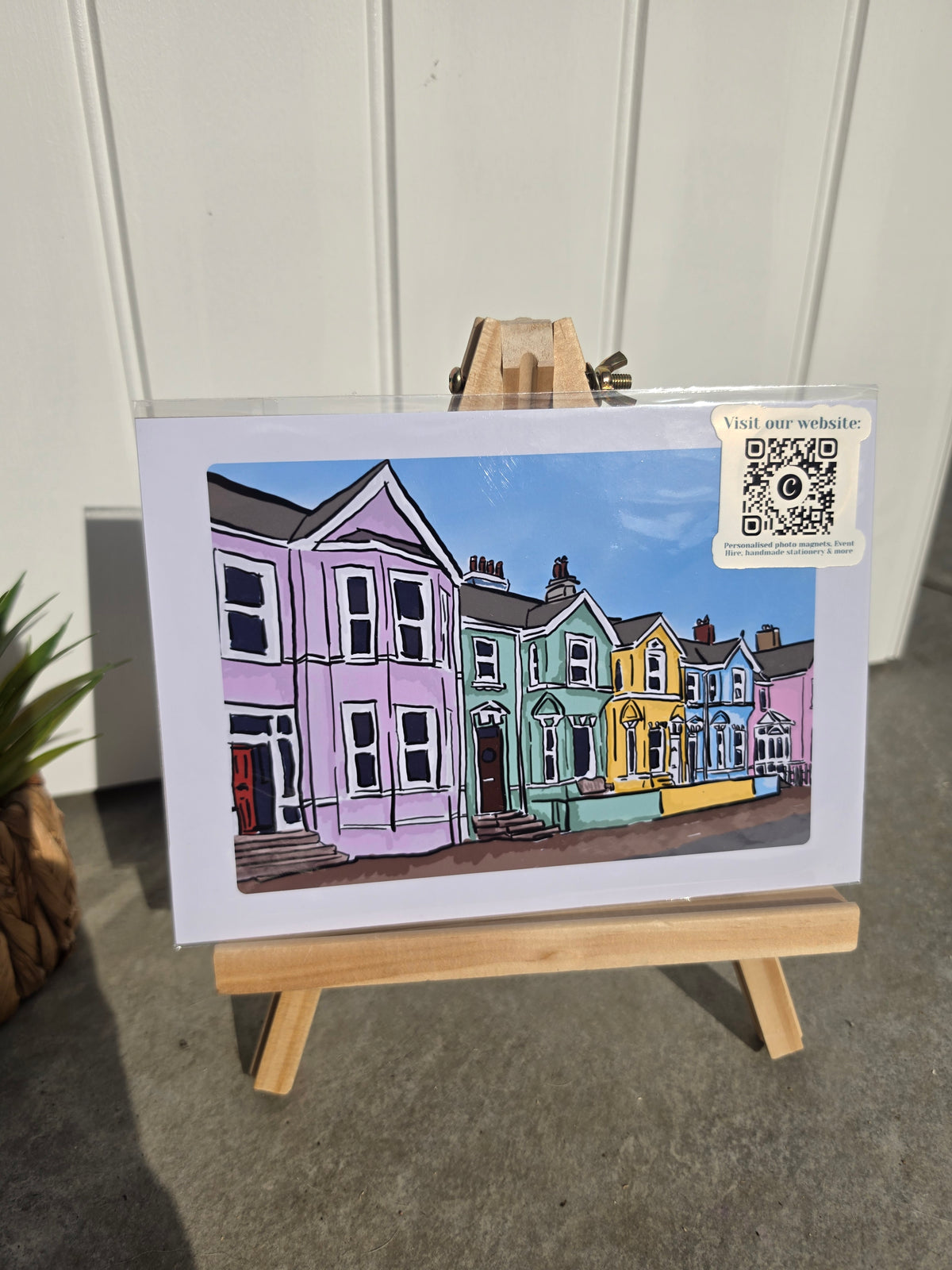 art print displayed on a small wooden easel, showing a colorful illustration of row houses in pastel pink, green, yellow, and blue. The print has a white border 