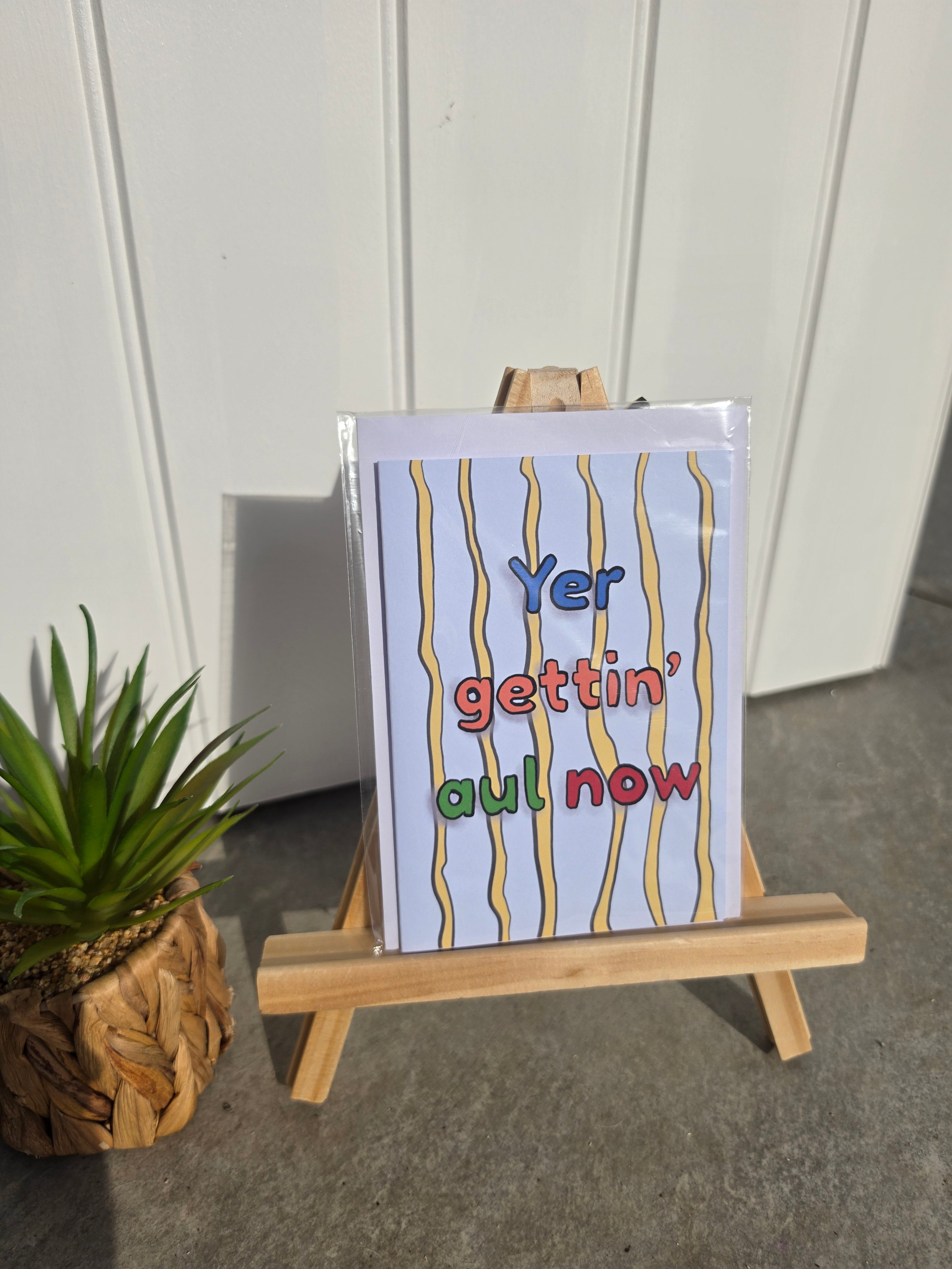 A greeting card with the phrase 'Yer gettin' aul now' in multicolored letters, displayed on a small wooden easel. The card has a white background with vertical yellow wavy lines and is photographed outdoors against a white paneled wall, with a small potted succulent to the left.