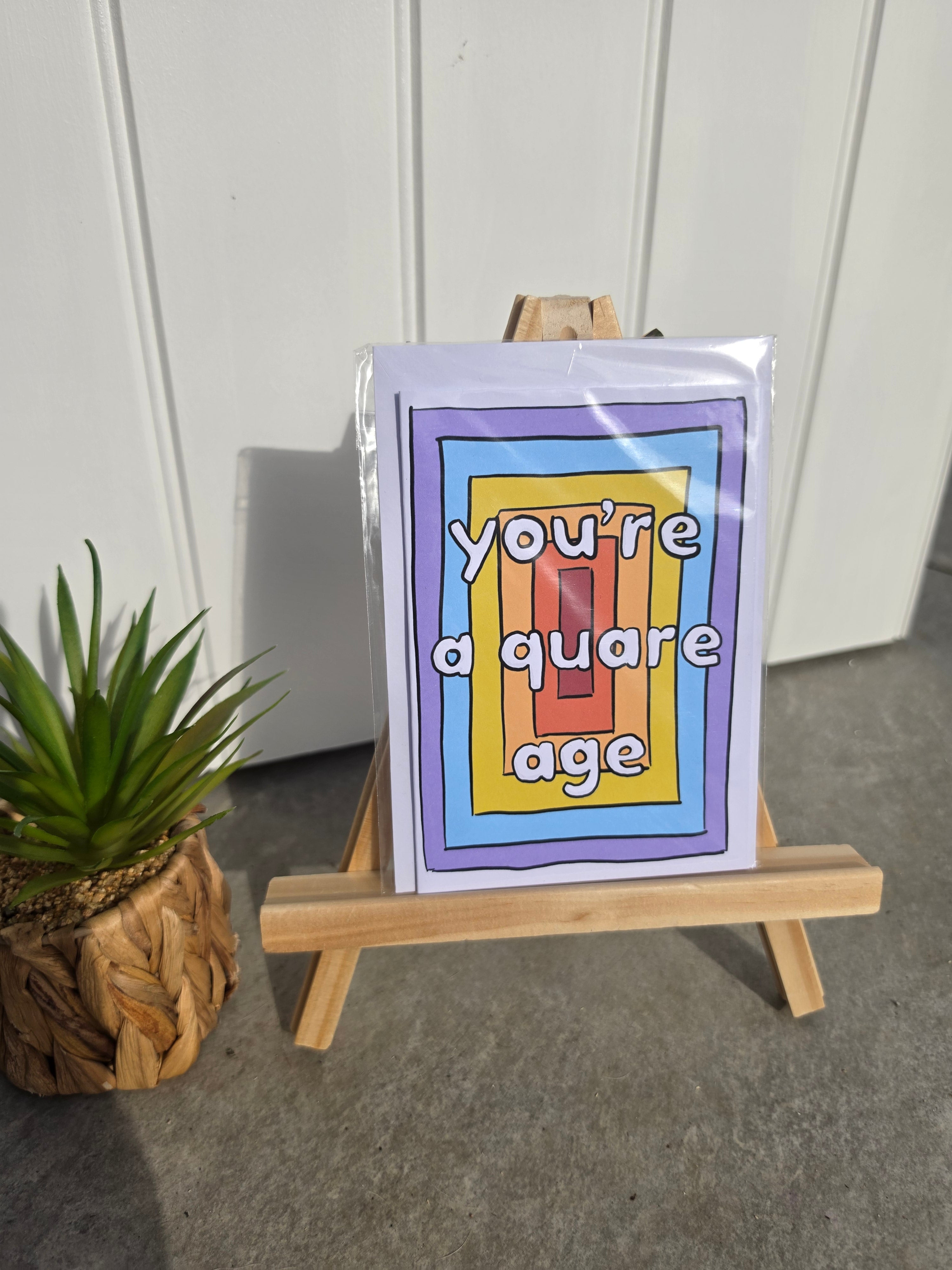 A square greeting card with a colorful geometric border and the text 'you're a quare age' displayed on a small wooden easel. The card is wrapped in clear plastic, set on a gray concrete floor next to a potted green plant against a white paneled wall.