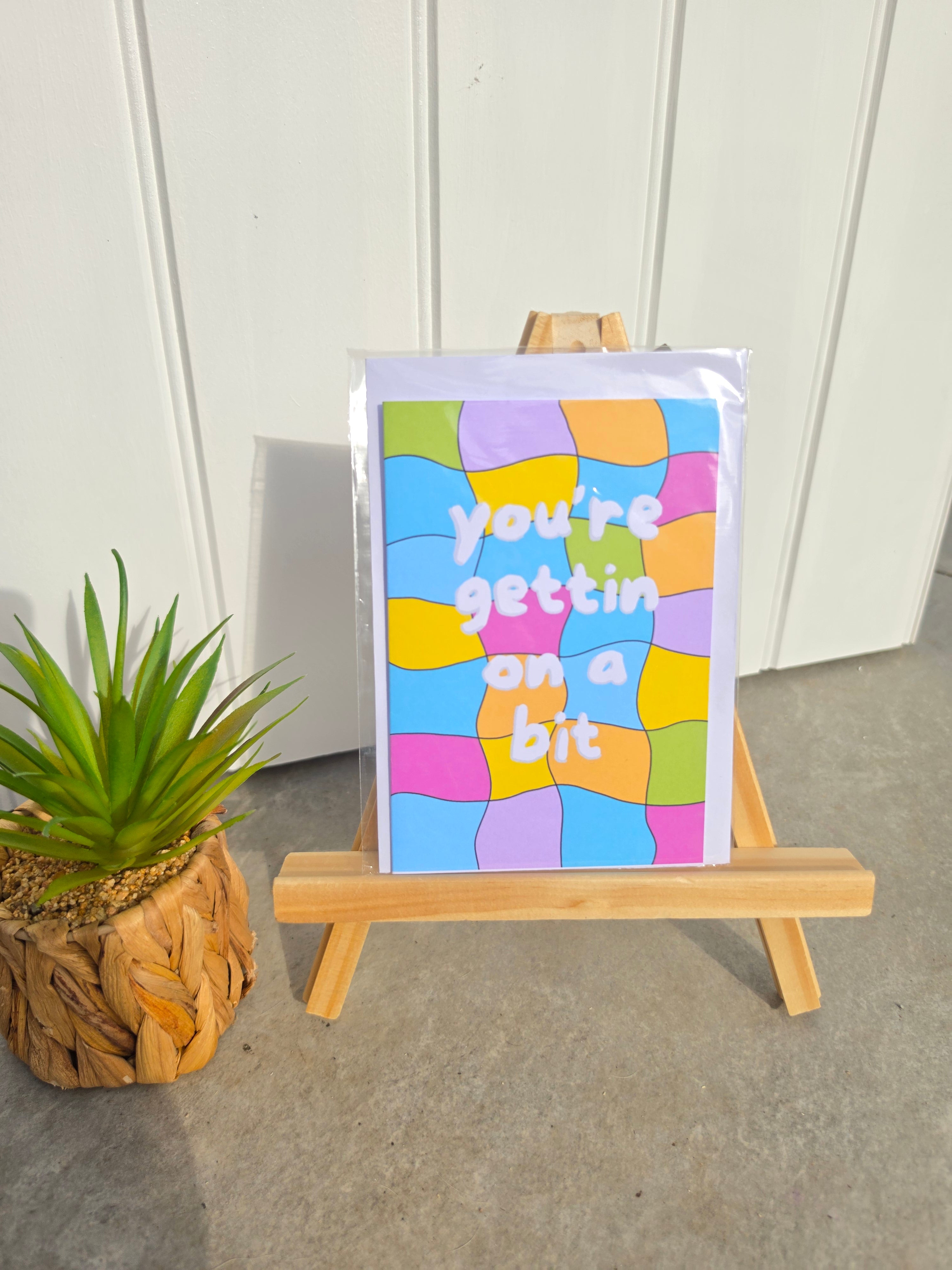 A colorful card with the text 'you're gettin' on a bit' in white lettering over a wavy, multicolored mosaic pattern, displayed on a small wooden easel next to a potted green plant.