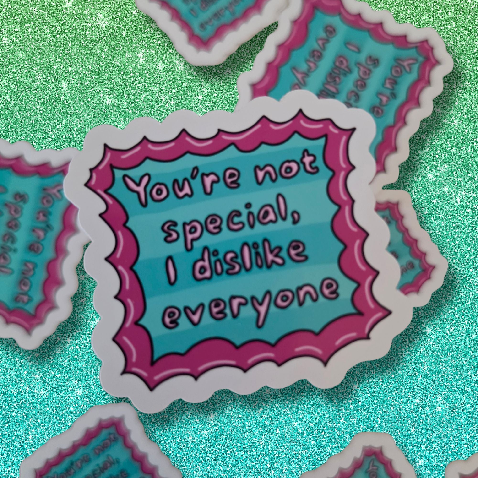 A close-up of a die-cut sticker with a scalloped border and the text 'You're not special, I dislike everyone' in pink lettering on a turquoise background with pink stripes. Other identical stickers are partially visible around it on a glittery turquoise surface.