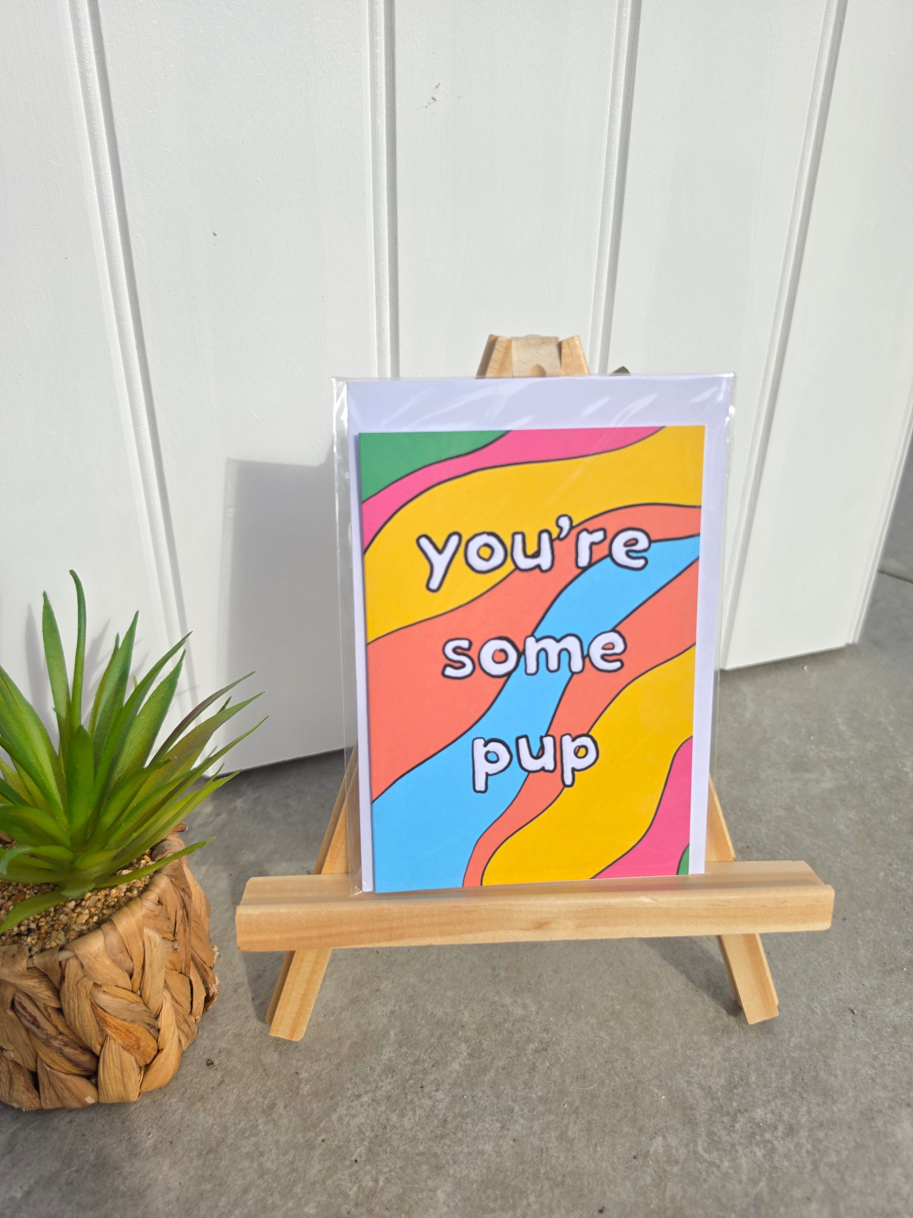 A colorful card with wavy stripes and the text 'you're some pup' displayed on a small wooden easel, with a potted succulent to the left against a white paneled wall.