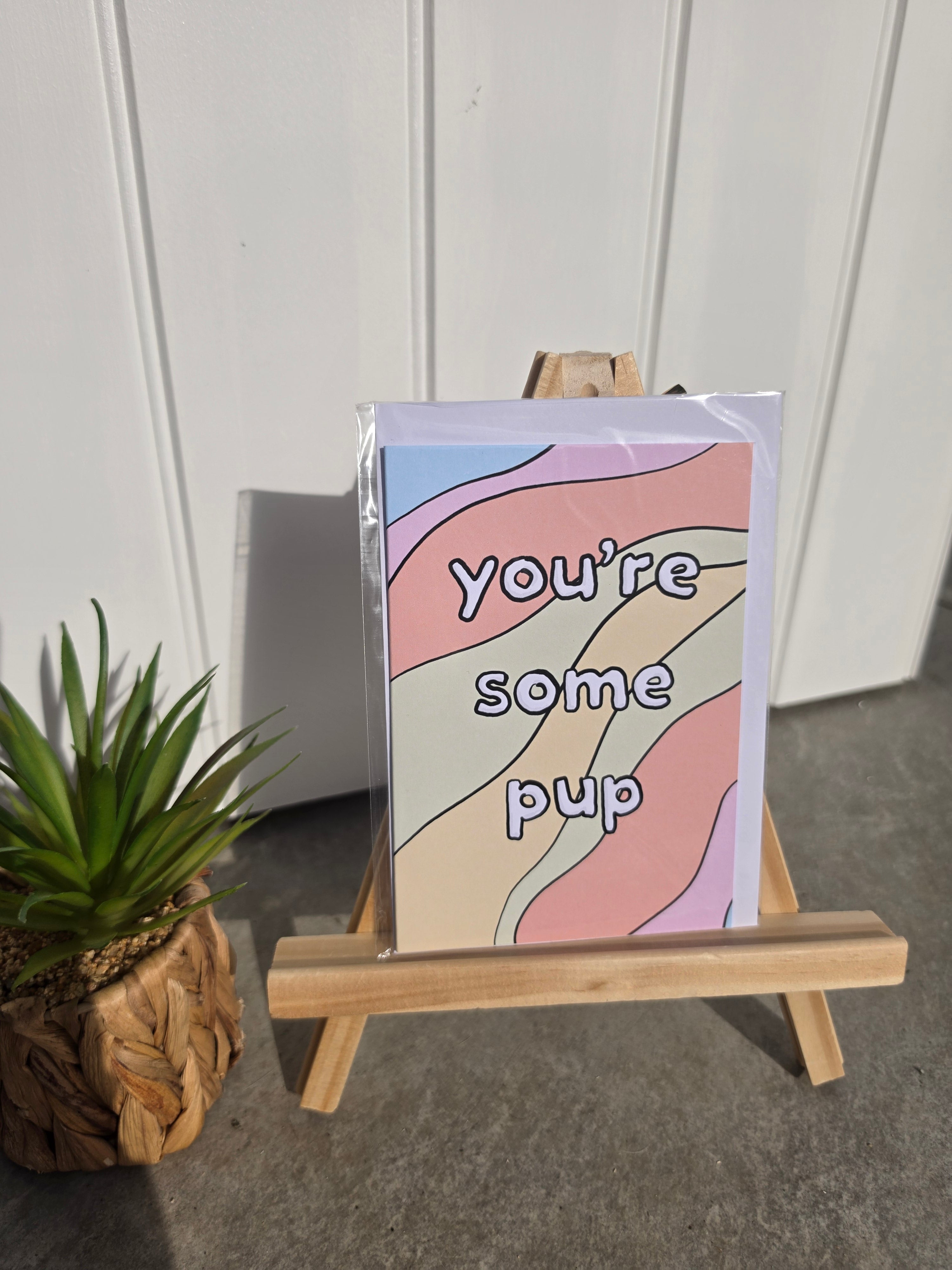 A greeting card with a pastel wavy pattern and the text 'you're some pup' in black outline lettering, displayed on a small wooden easel next to a potted succulent.