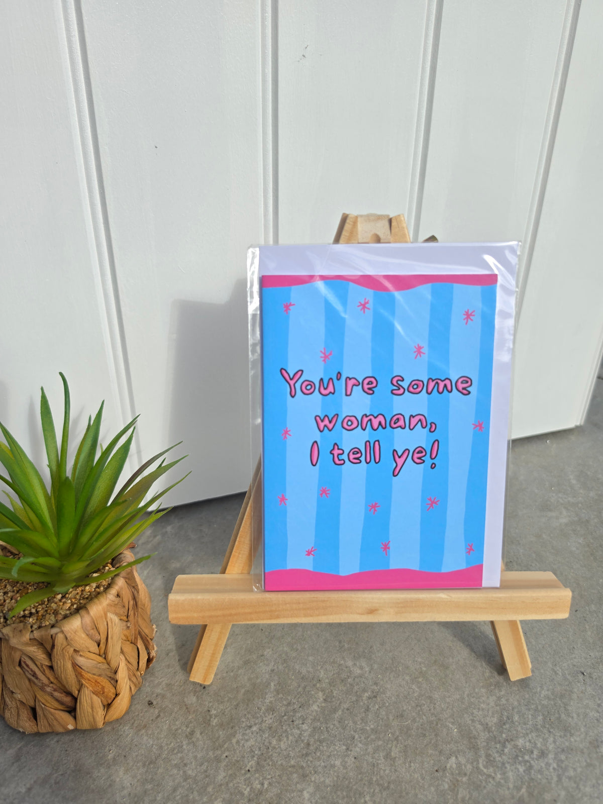 A greeting card with a blue and pink striped design and the text 'You're some woman, I tell ye!' displayed on a small wooden easel next to a potted plant.